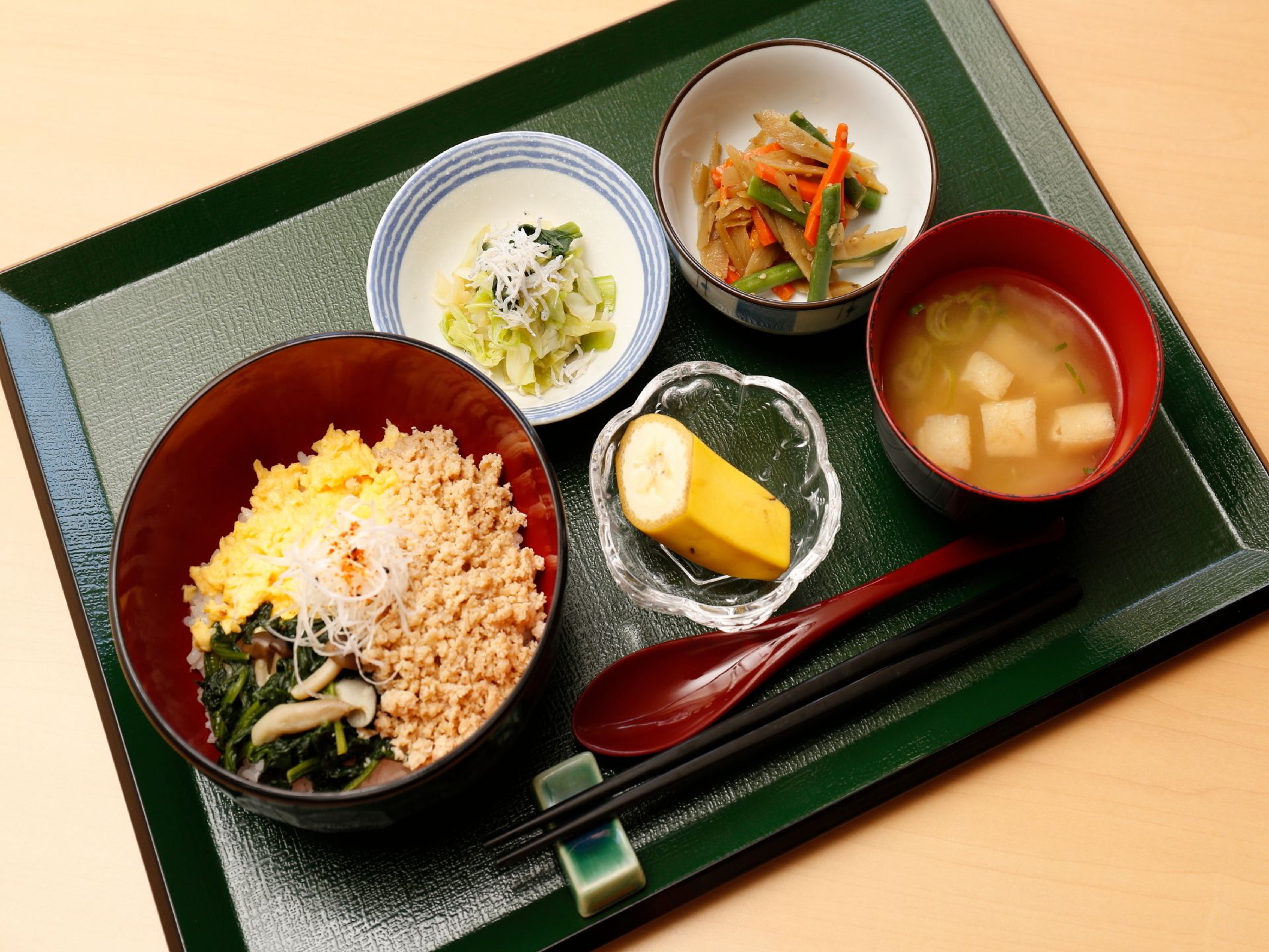 長方形のトレーに1人分の食事がセットされている。手前には箸とスプーンが置かれ、果物と味噌汁を含む5品が並んでいる。