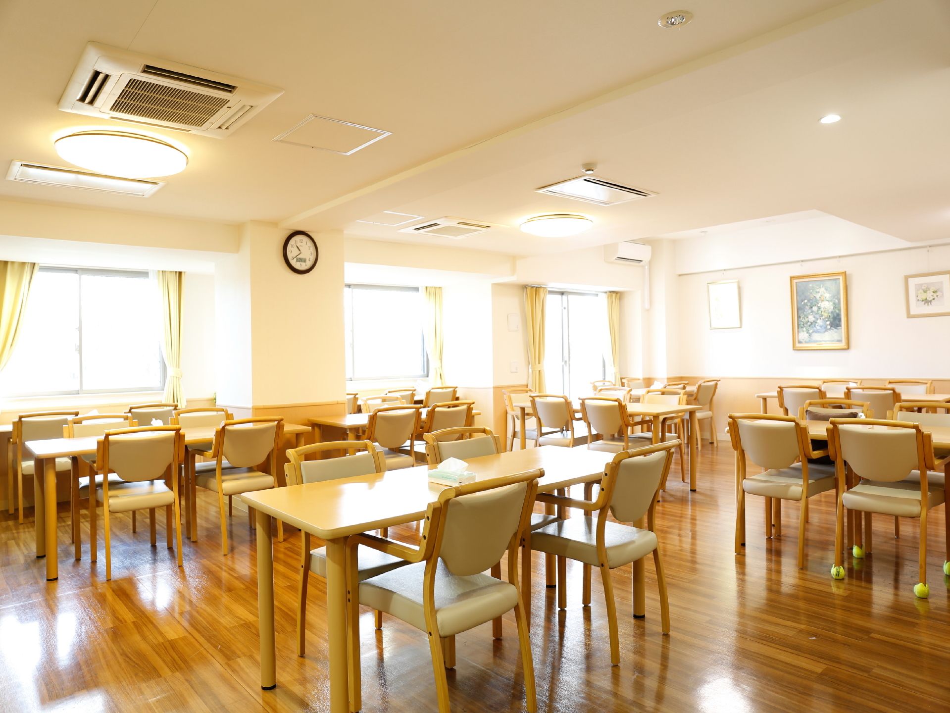 黄色いカーテンが取り付けられている大きな窓3か所からは自然光が入り、明るい。テーブルや椅子がいくつも並べられている。