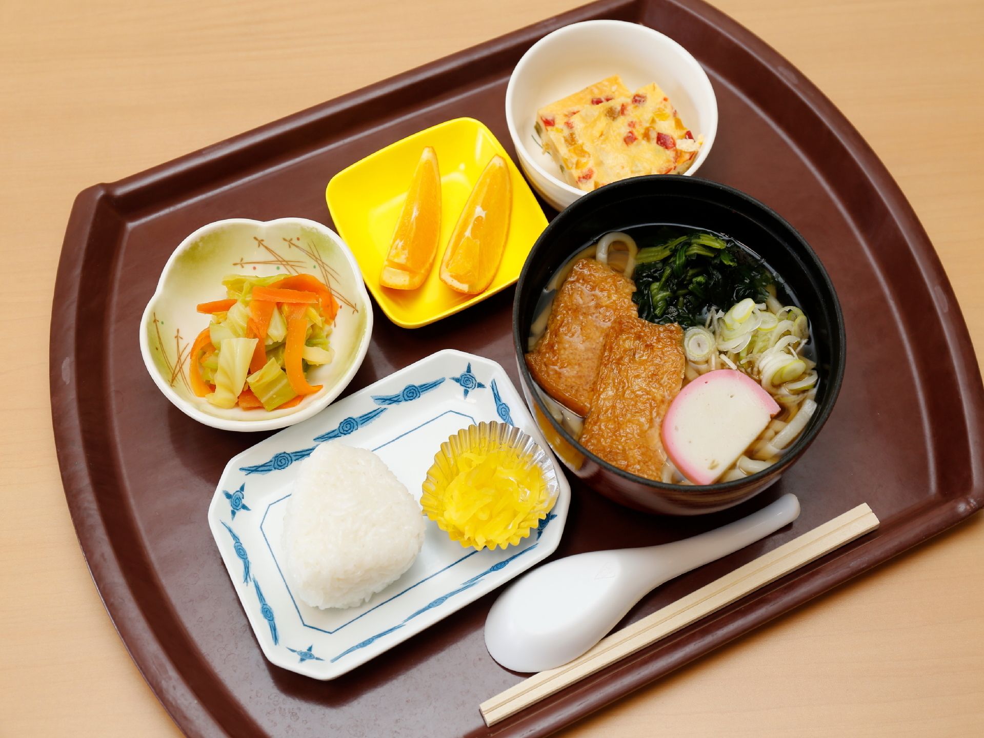 食事が5品用意されている。油揚げとかまぼこ、ネギ、若芽が乗ったうどんや漬物付のおにぎり、ニンジンとキャベツを使ったおかずなどがある。