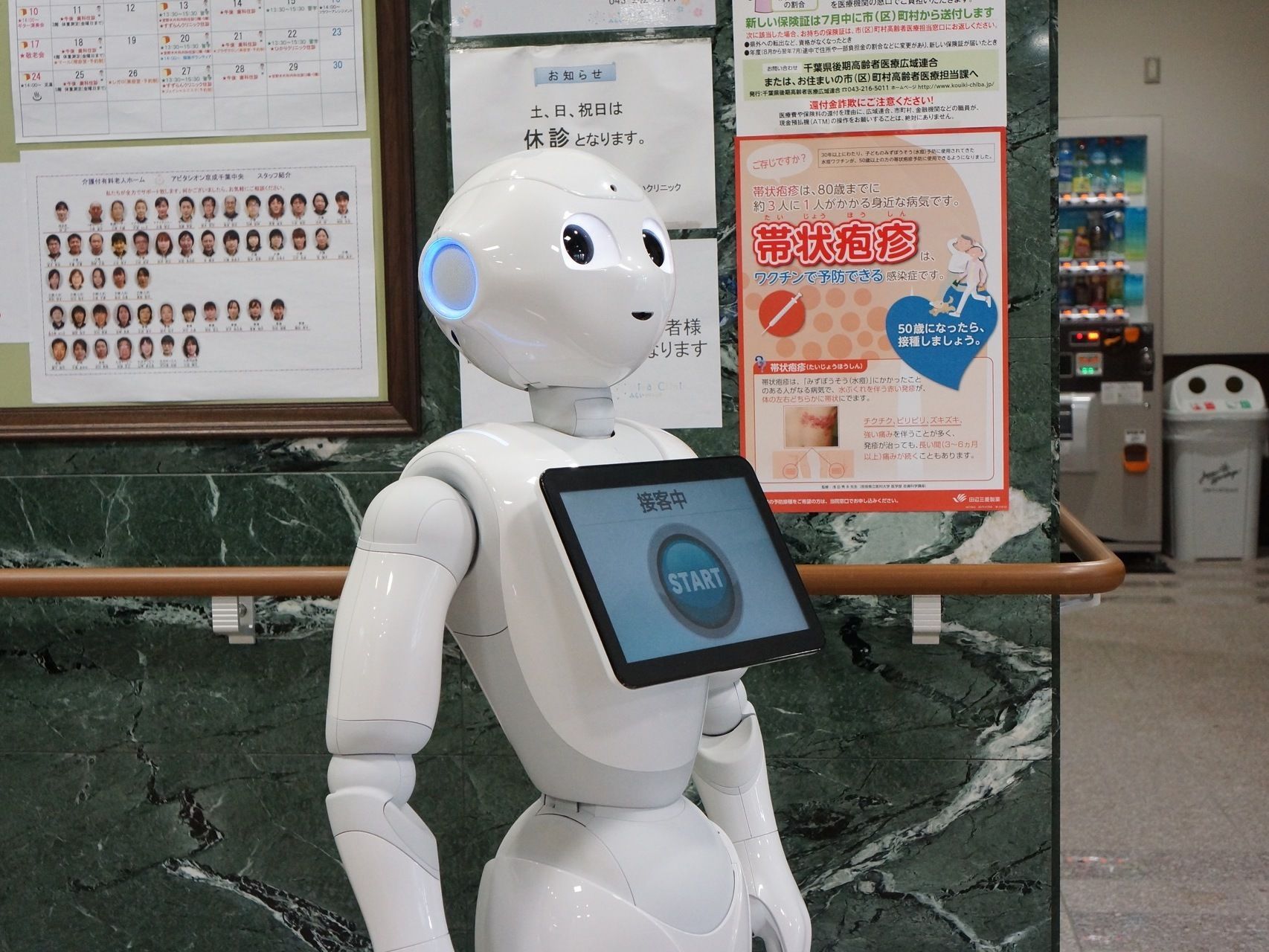 受付があるロビーの壁面には掲示物が貼られている。白色の人型のロボットがあり、その胸部に備わっているタブレットに接客中と表示されている。