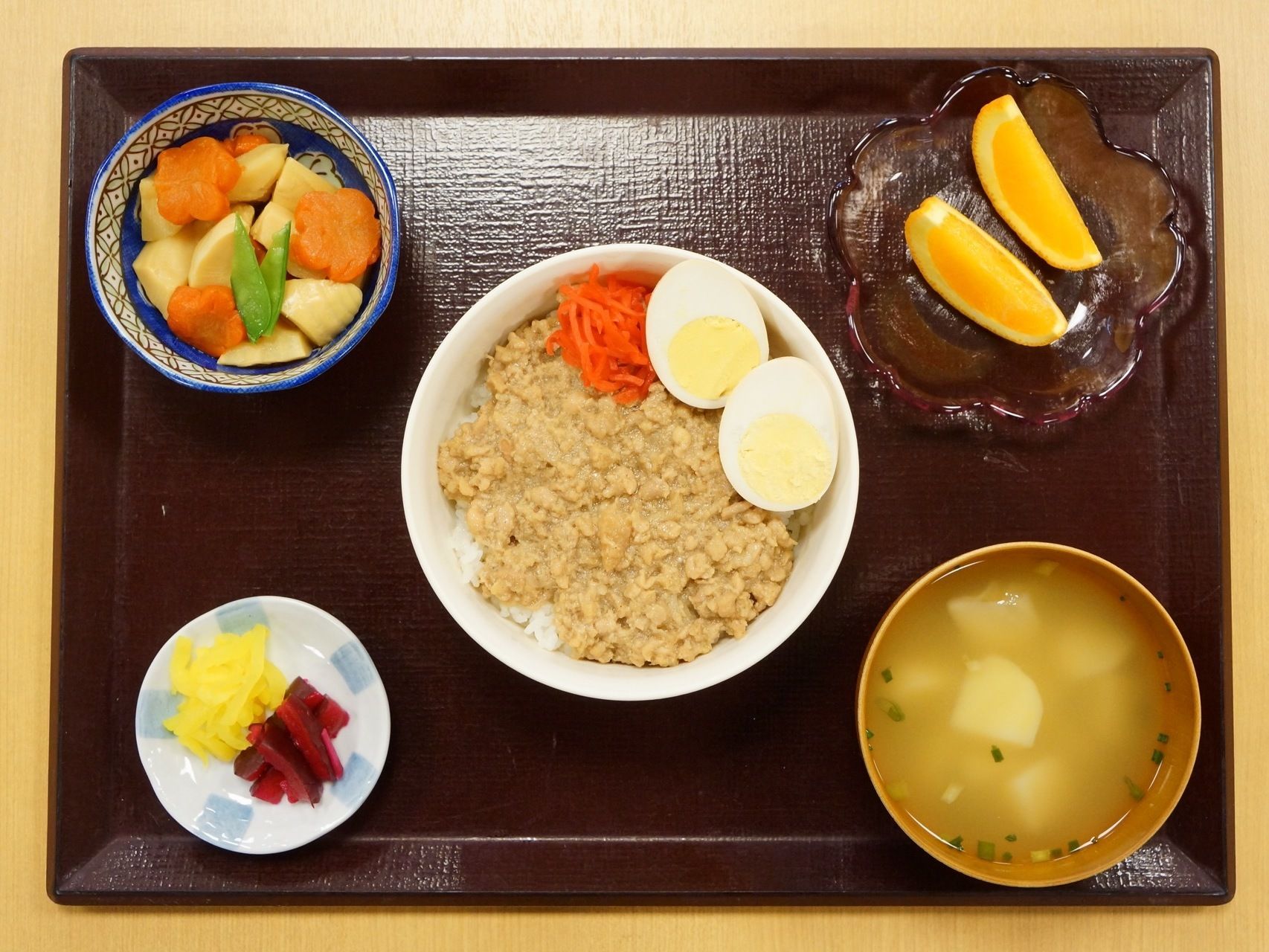 食器に盛り付けられて食事が茶色のお盆の上に並べられている。根菜の煮物には、人参が花形に飾り切りされて添えられている。