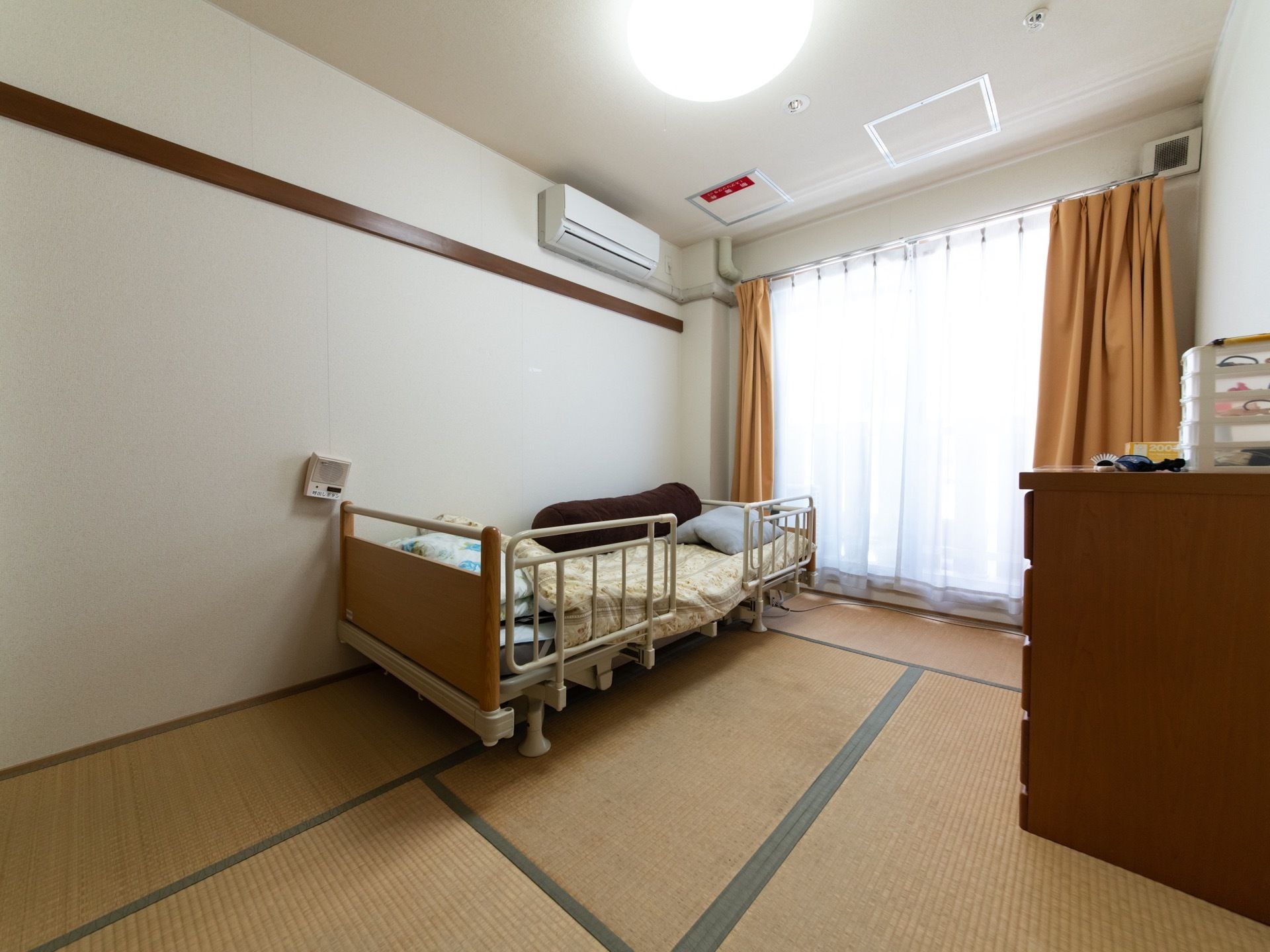 茶色を基調とした畳敷きの色合いの室内には、大きな窓から日差しが注ぎ込んでいる。設置されたベッドには、転落防止用の柵が取り付けられている。