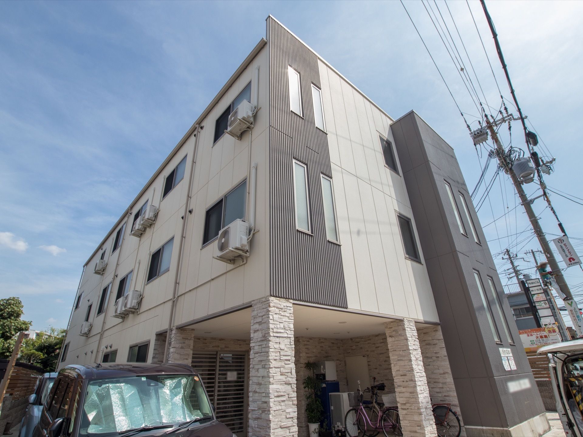 外壁がグレーとホワイトで施された、おしゃれな雰囲気の建物である。1階の屋根下に自転車が数台停められている。