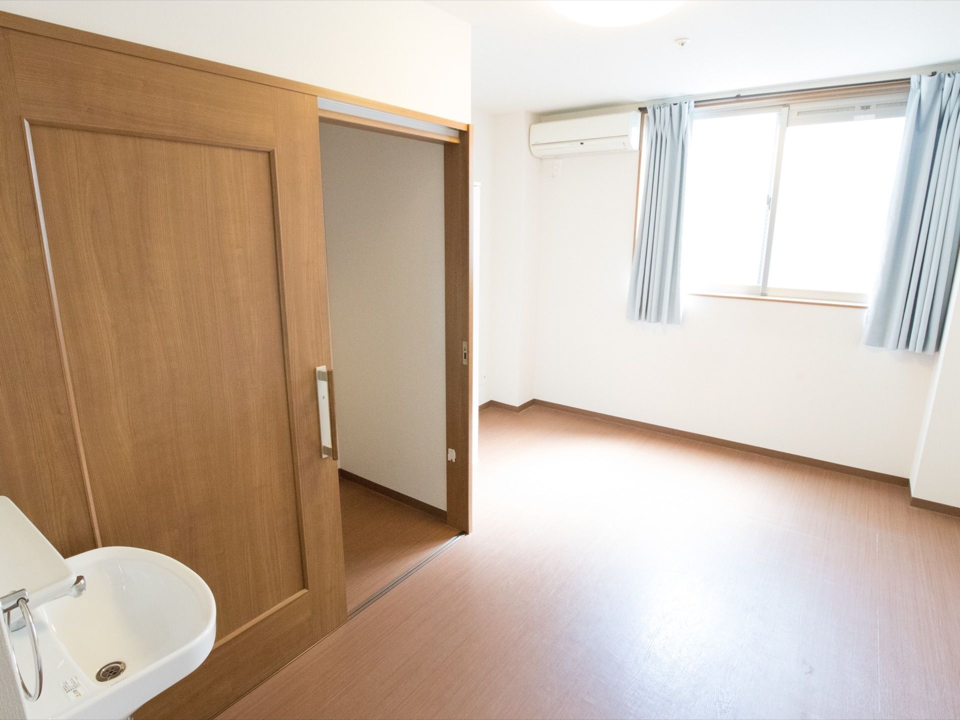 窓に淡いブルーのカーテンが設置されている。そばにはエアコンを完備。広々とした建具が備えつけられている。