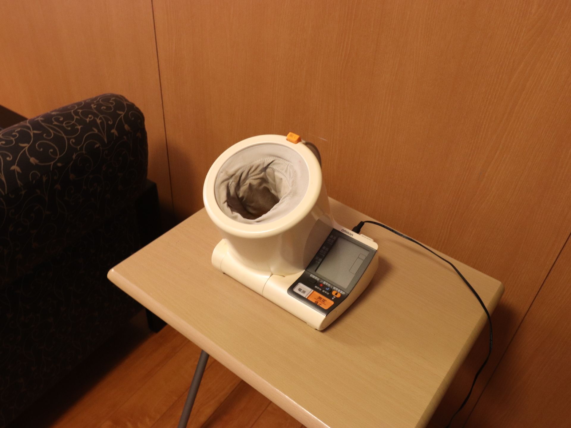 フローリングの部屋の壁際に木製のテーブルが置かれており、上部に自動血圧計が設置されている。左側にはソファーがある。