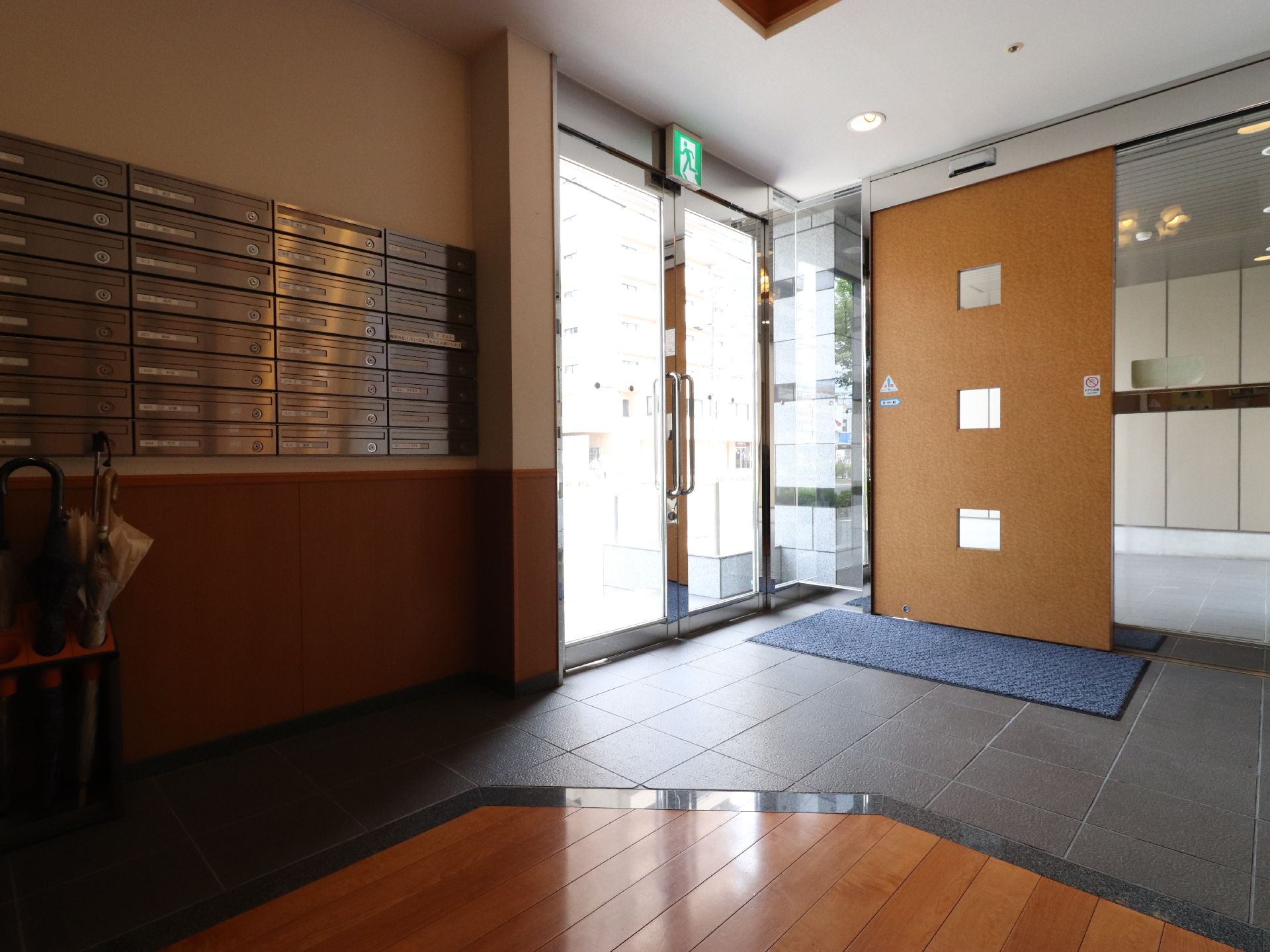ガラス張りの両開きのドアと木目仕様の自動ドアが垂直に設置されている。奥の壁際には郵便受けが並んでいる。ダークグレイのタイル調の上り口である。