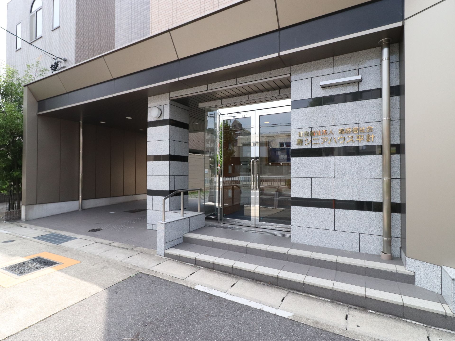 薄いグレイ色と黒色のボーダーの外壁にガラス張りの玄関ドアのエントランスである。外壁には施設名が施されている。ドア前は2段階段になっている。