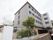 施設の看板は、白がベースで文字が緑色のすっきりとしたデザインである。建物は４階建てで、外壁の色はグレーである。