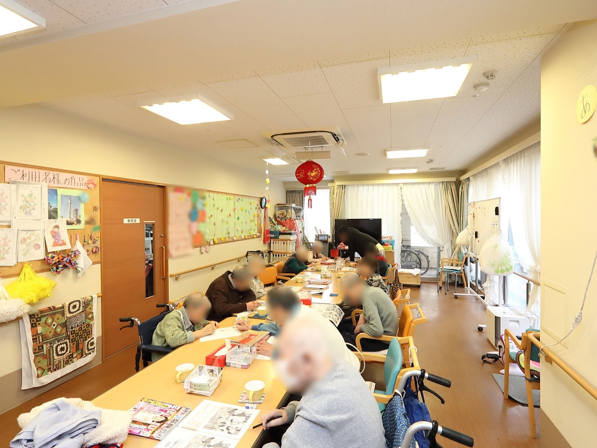 縦長の広々とした娯楽室は角部屋なので窓がたくさんあり、日射しが差し込み常に明るい。テーブルが一列に並べられている。