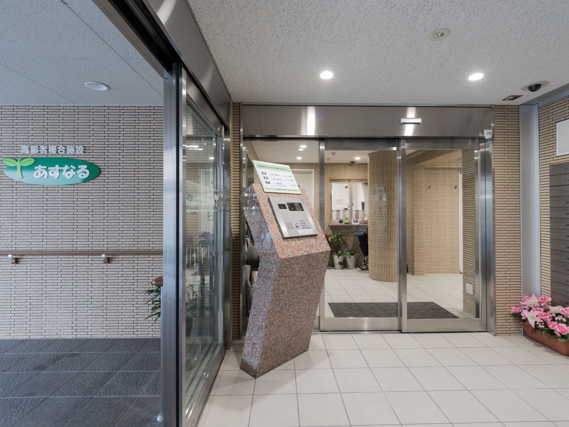 施設入口の外壁には施設名が書かれた看板が貼り付けられている。手すりも取り付けられている。中に入るとオートロックの操作ボタンがある。