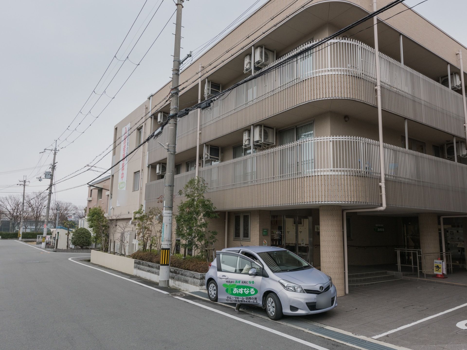 施設は３階建てである。建物の前には施設専用の送迎車が１台停められている。車には施設名が書かれている。