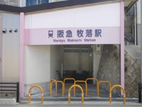 黄色の逆Ｕ字アーチ型車止が並んだ上部に、ピンクの板に青文字で阪急牧落駅と大きく書かれた看板が掲げられている。