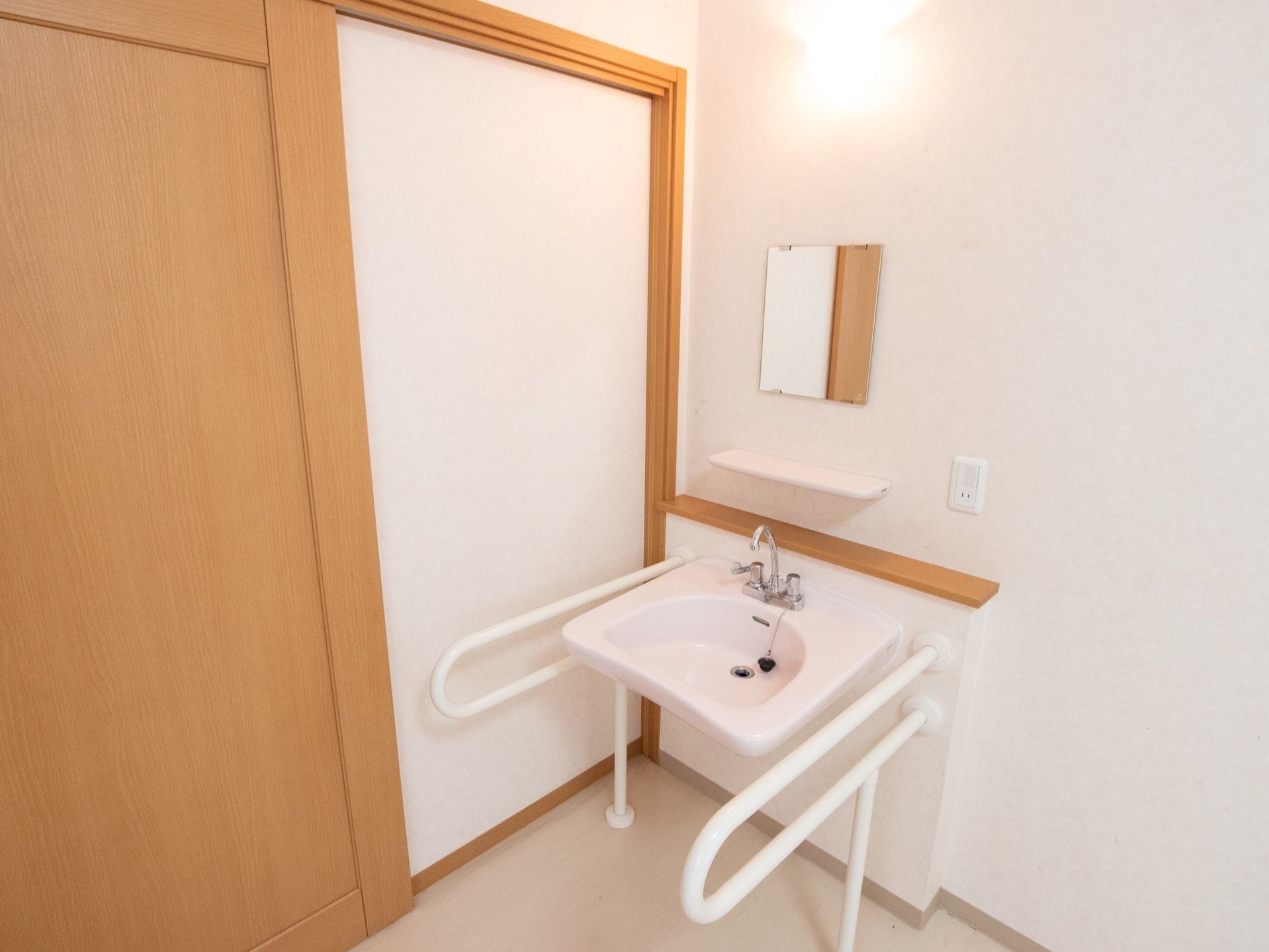 居室には、両サイドに手すりが設置された洗面台が完備されている。鏡や棚、コンセントが壁に取りつけられている。