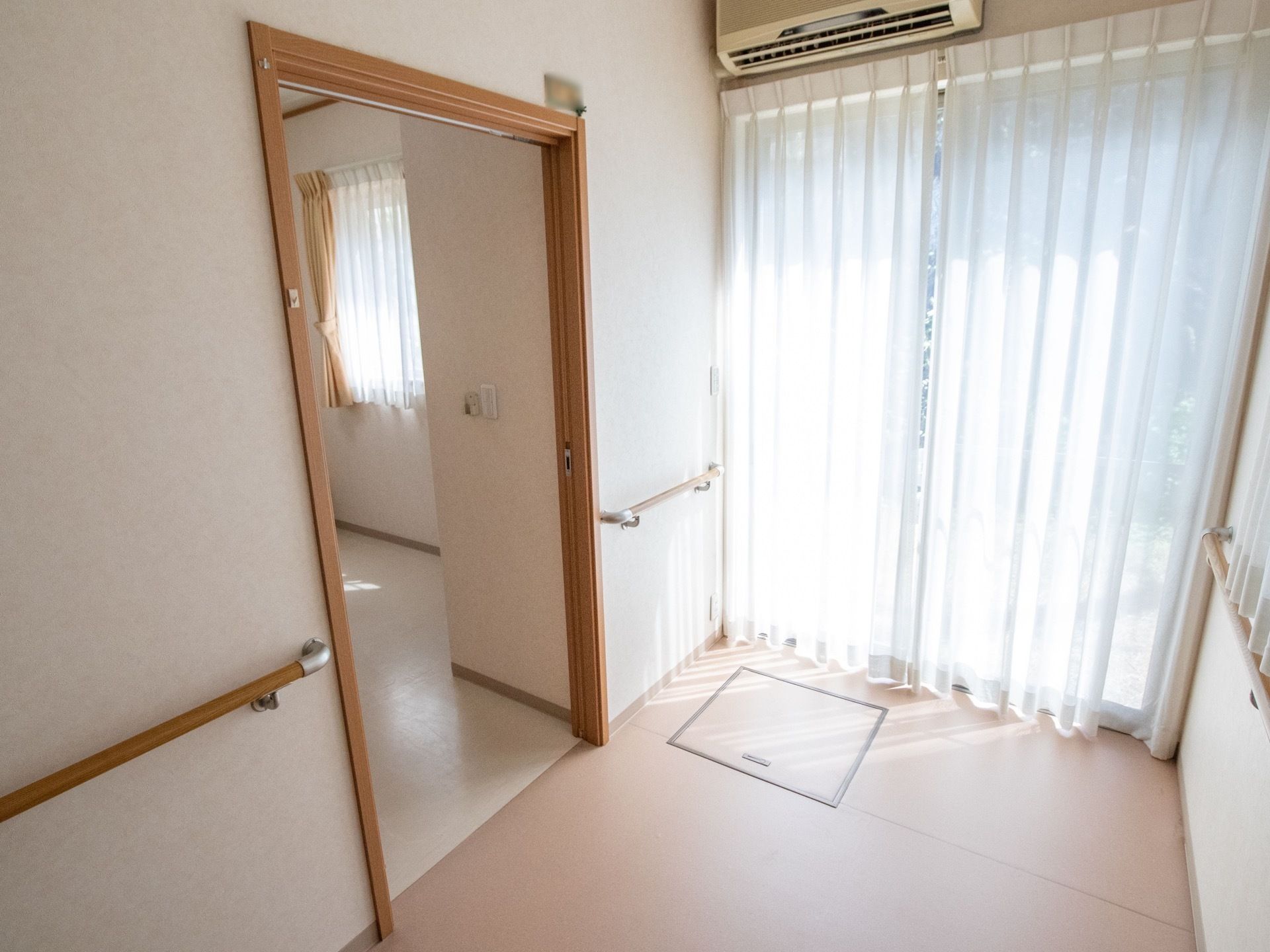 廊下の突き当りには、レースのカーテンが掛けられた窓がある。明るい日差しが差しこんでいる。エアコンや手すりが壁に設置されている。