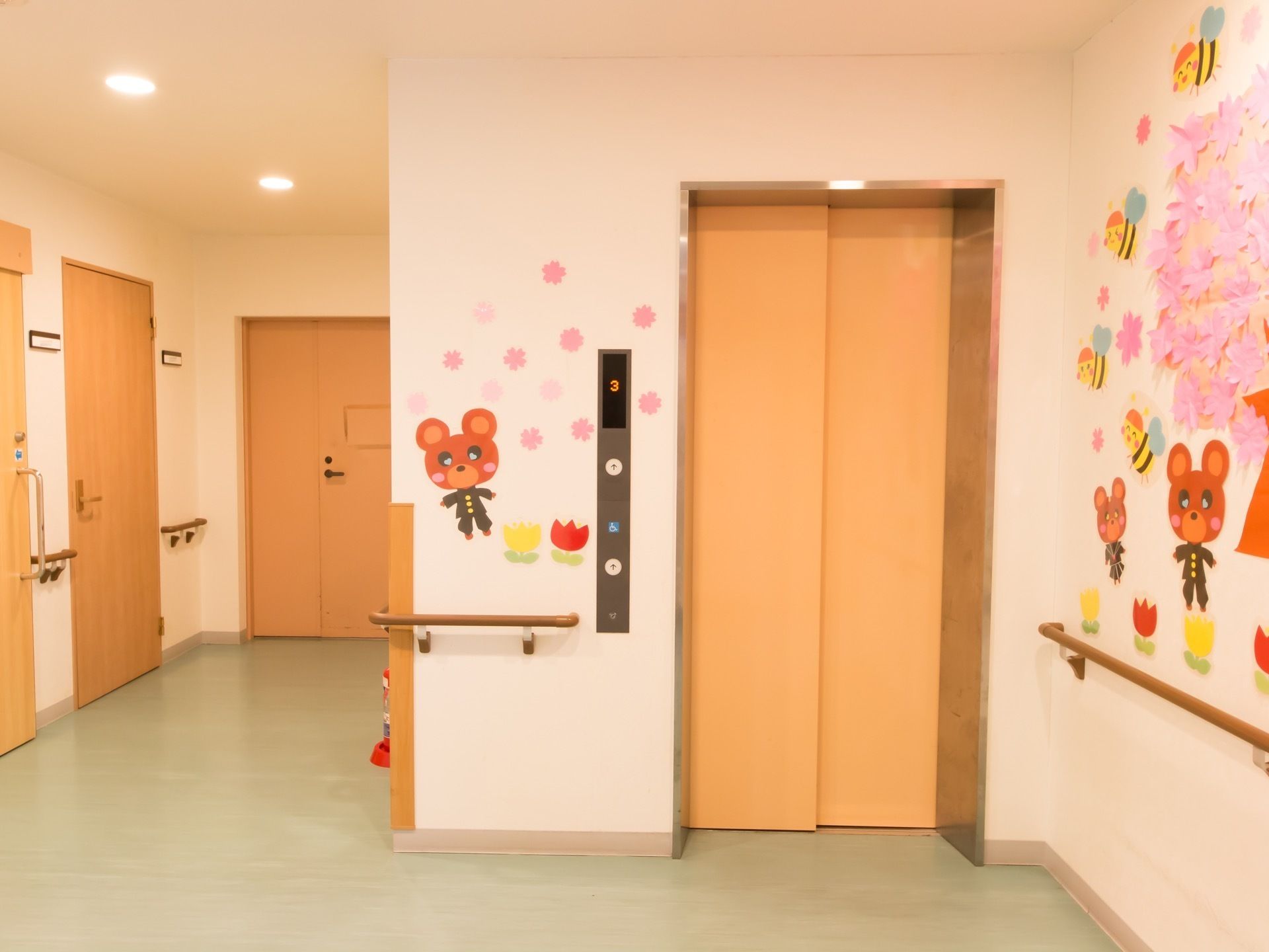 クリーム色の扉のエレベーターが1基ある。エレベーター横の壁には桜の木と蜂、学生服を着たクマのイラストが貼られている。