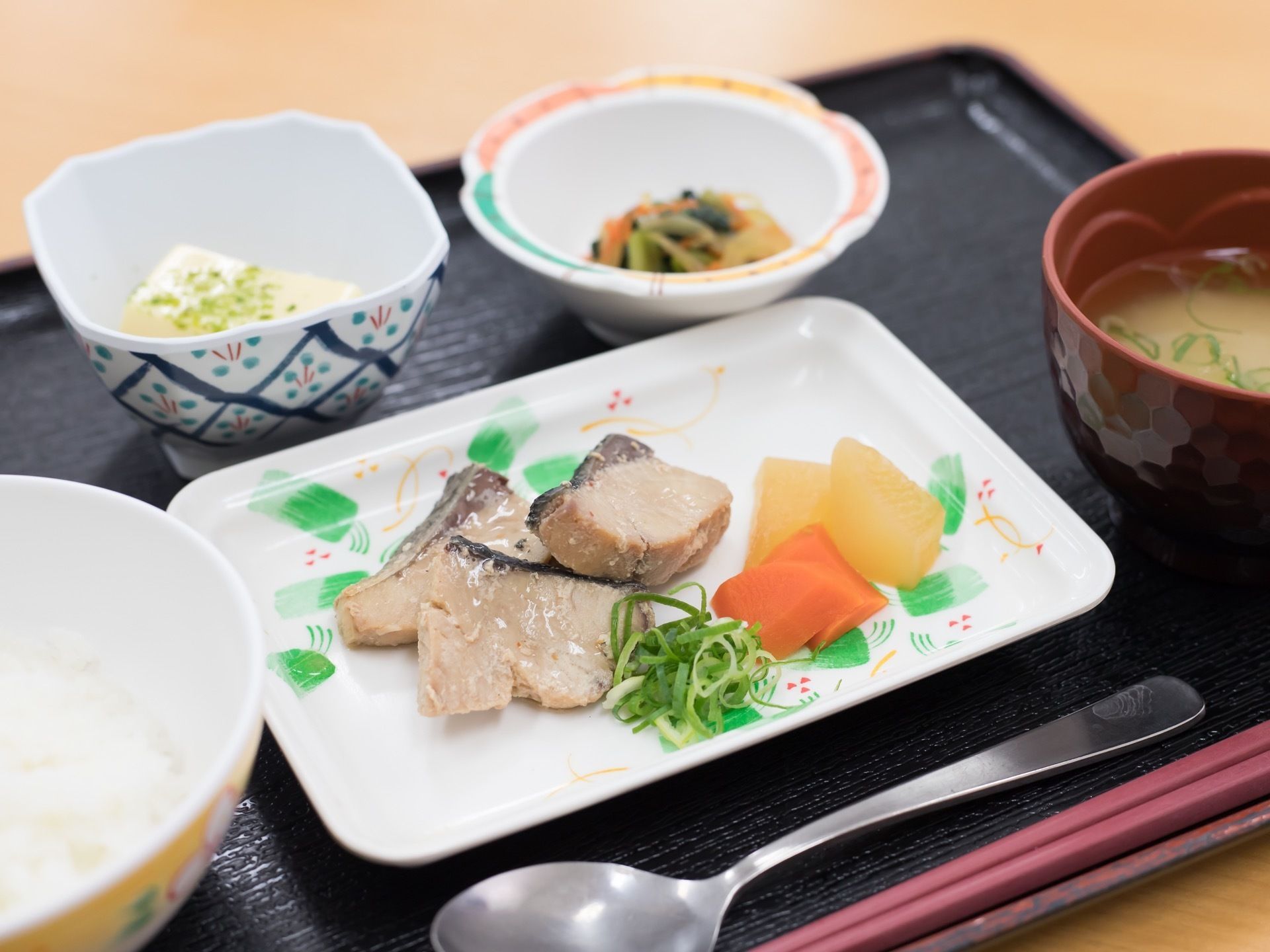 黒いお盆の上に食事が配膳されている。ごはん、お味噌汁、おかずが3品のメニューである。箸、スプーンも置かれている。