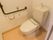 トイレには洋式便器が設置されている。壁側には茶色いL字状の手すり、呼び出しボタン、トイレットペーパーフォルダーがある。