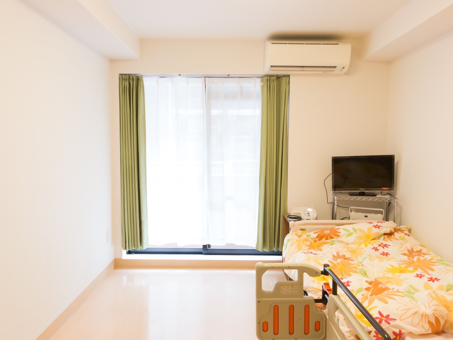 白を基調としたインテリアの居室である。介護ベッドが置かれ、大きな花柄の布団カバーが使われている。エアコンが備わっている。