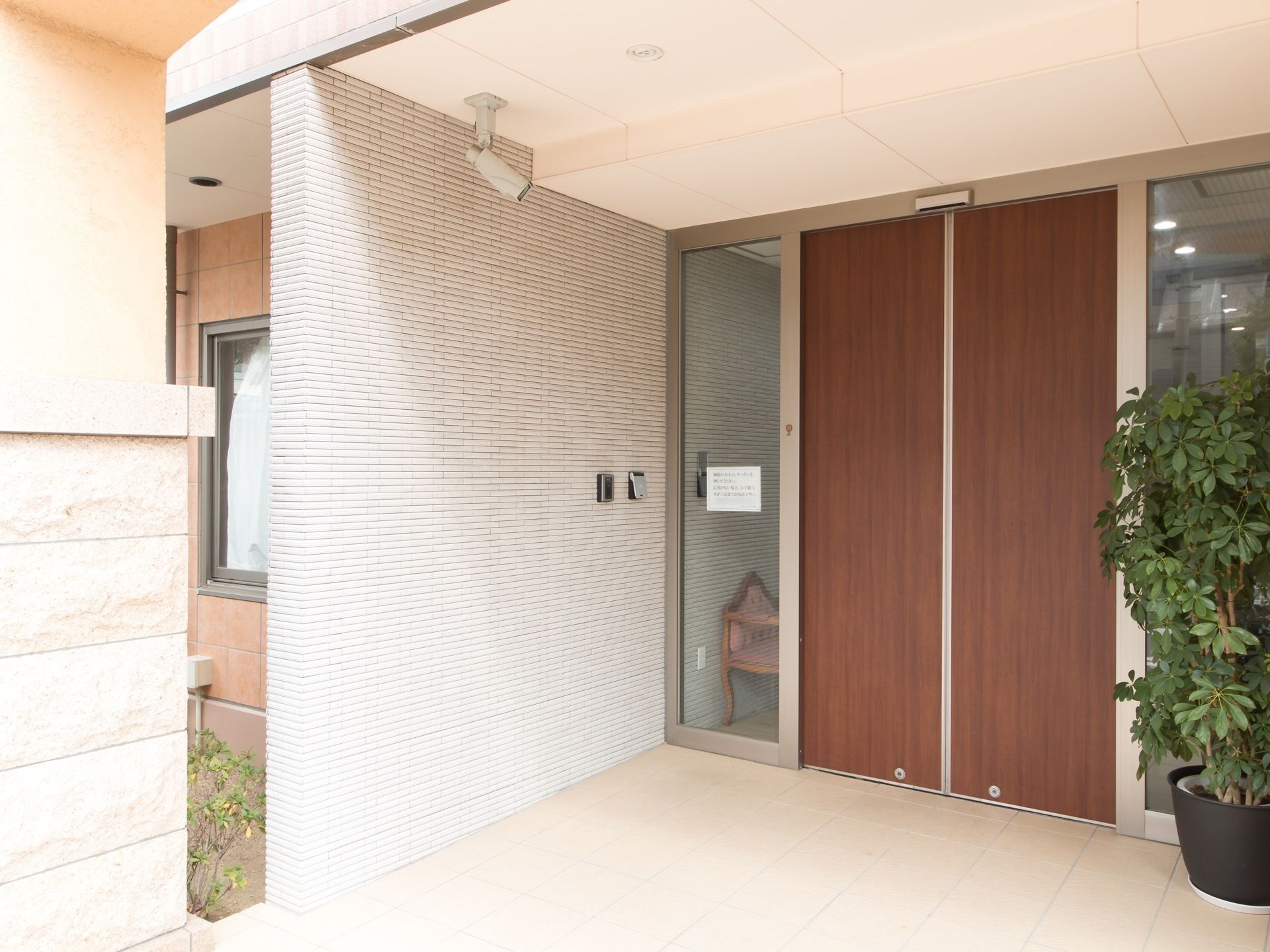 玄関は木目の美しい自動ドアで、ポーチには屋根がある。入口の手前に、大きな観葉植物の鉢植えが飾られている。