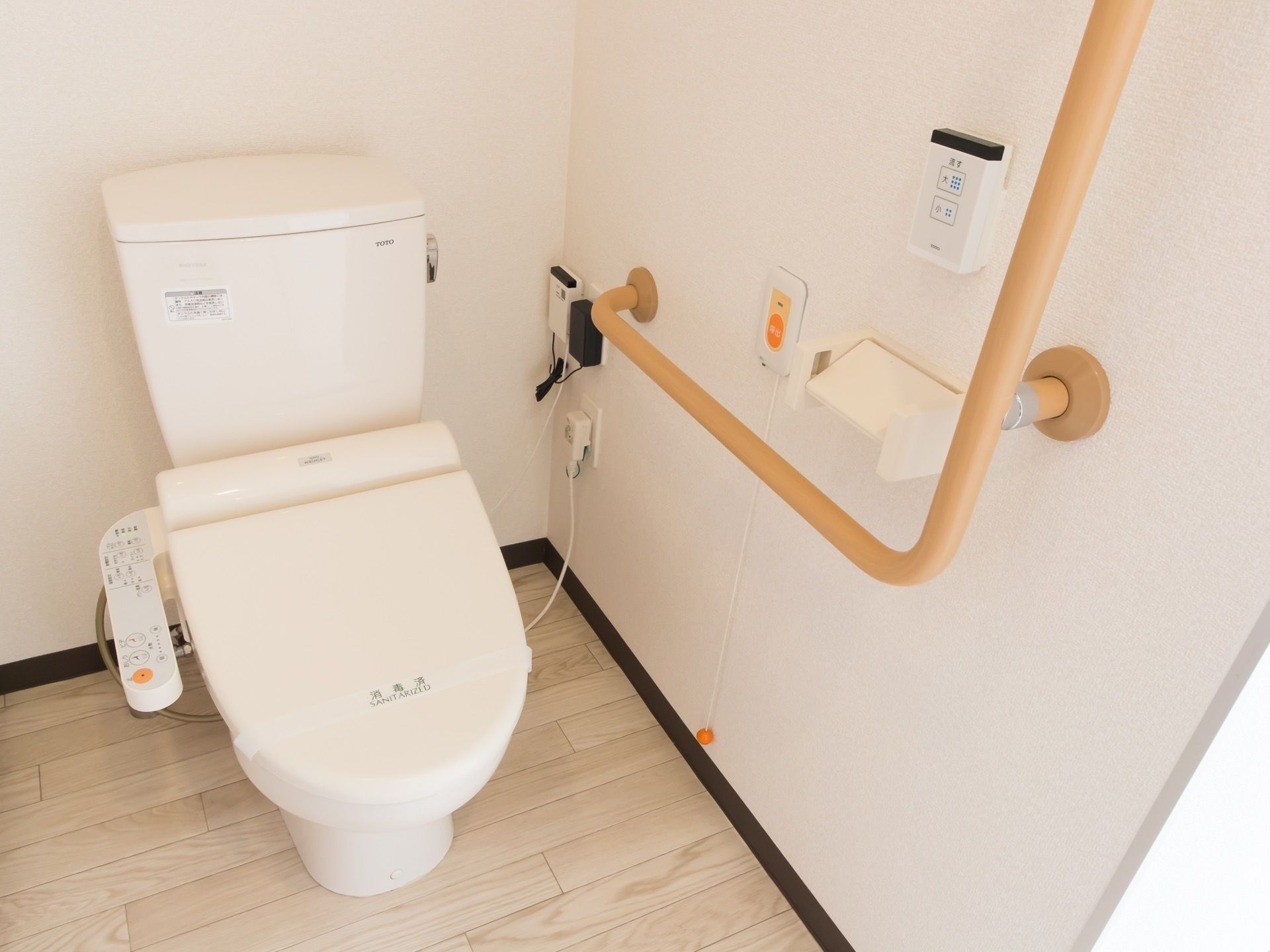 温水洗浄便座付きのトイレである。壁にはＬ字型の手すりや、ナースコール、トイレの洗浄ボタンが設置されている。