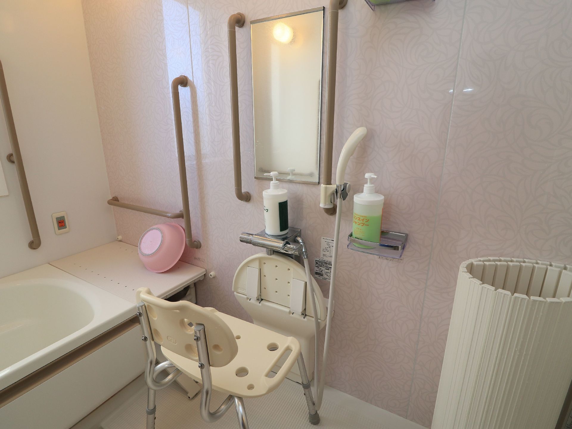 片面の壁は、淡いピンク色である。身体を洗う場所には、白色のシャワーチェアを完備。縦型の鏡も備えつけられている。