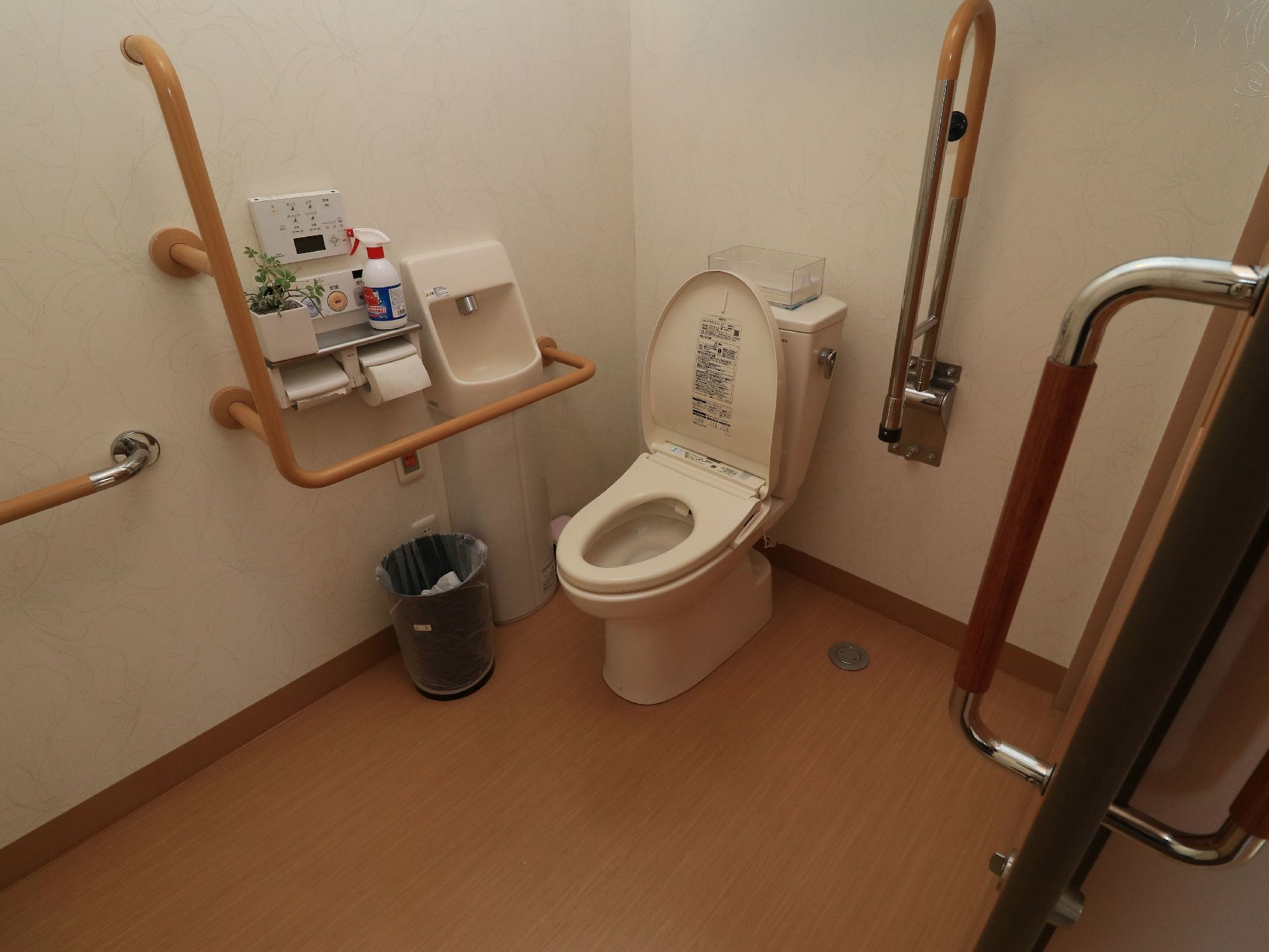広々としたトイレスペースに洋式便器と手洗いを設置している。壁には小物棚付きペーパーホルダーと複数の手すりを取りつけている。