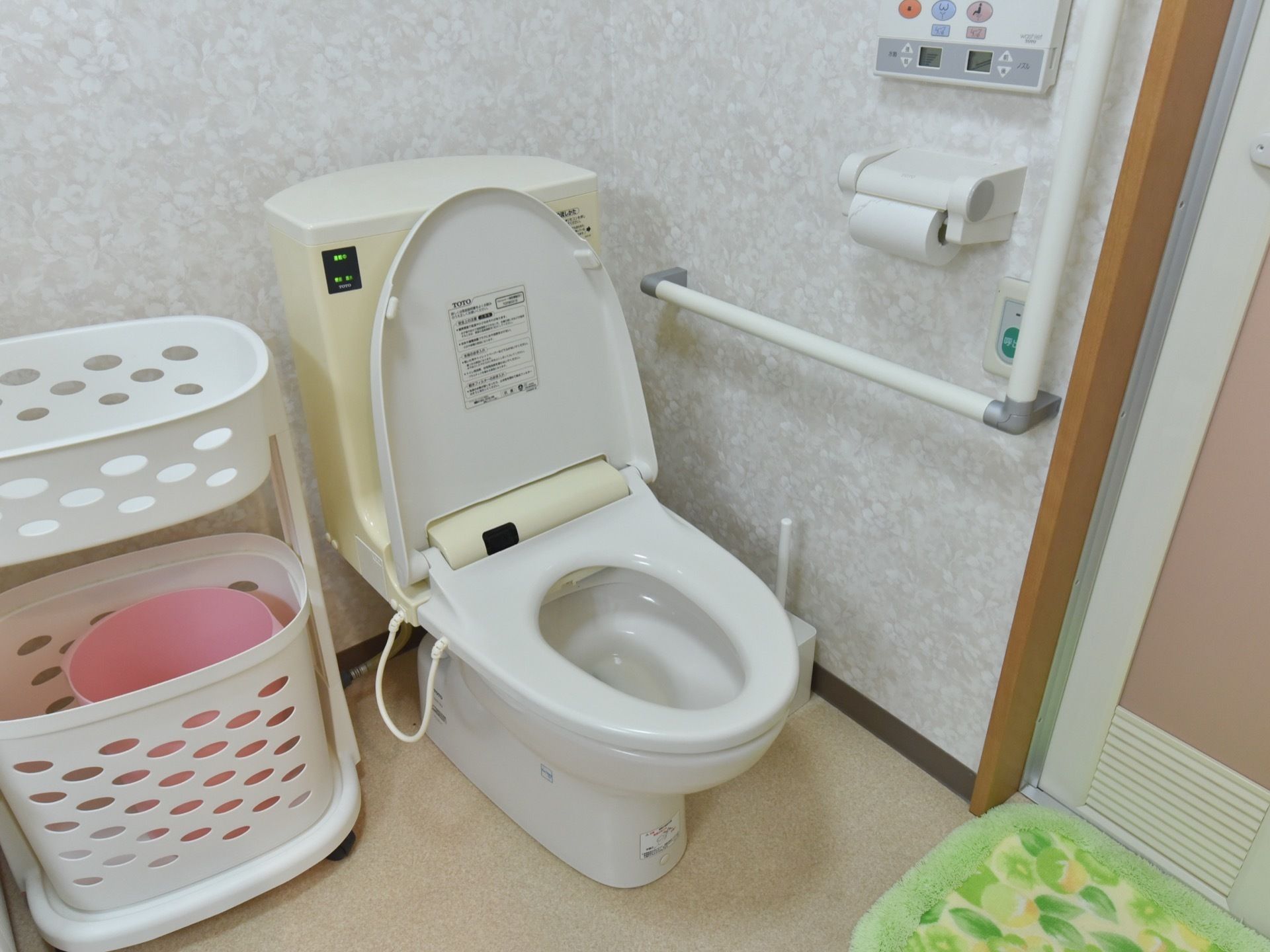 グレーを基調としたクロスが落ちついた雰囲気のトイレスペース。便器の横に洗濯かごが置かれている。壁には手すりと緊急通報装置が設置されている。