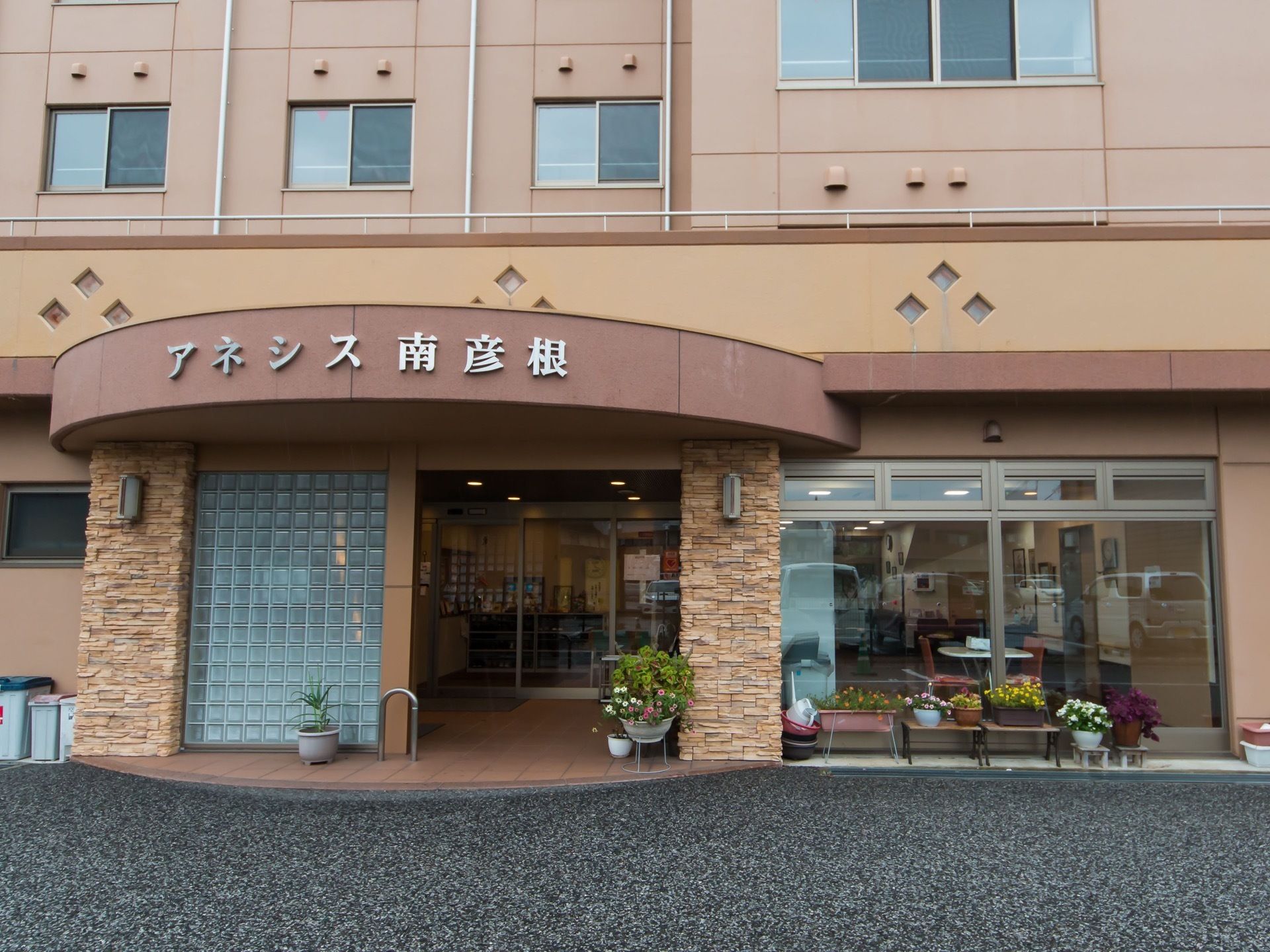 玄関は丸く張り出したえんじ色のひさしに施設名が大きく書かれている。周りには複数の植物が植えられた植木鉢が並べられている。