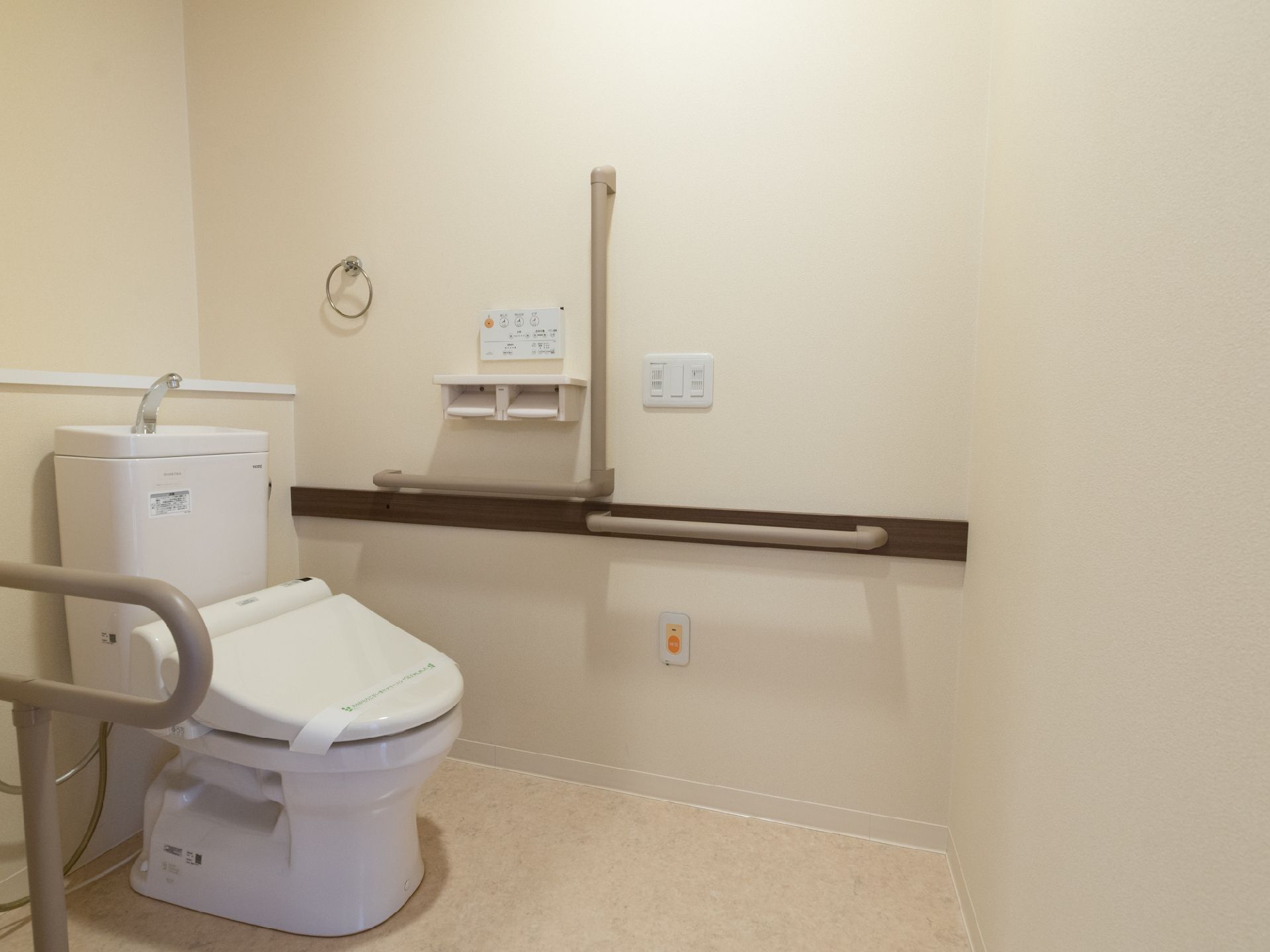 白色基調のトイレスペースである。便器の両側と前方の壁には手すりが設置されている。また、タオル掛けも取り付けてある。