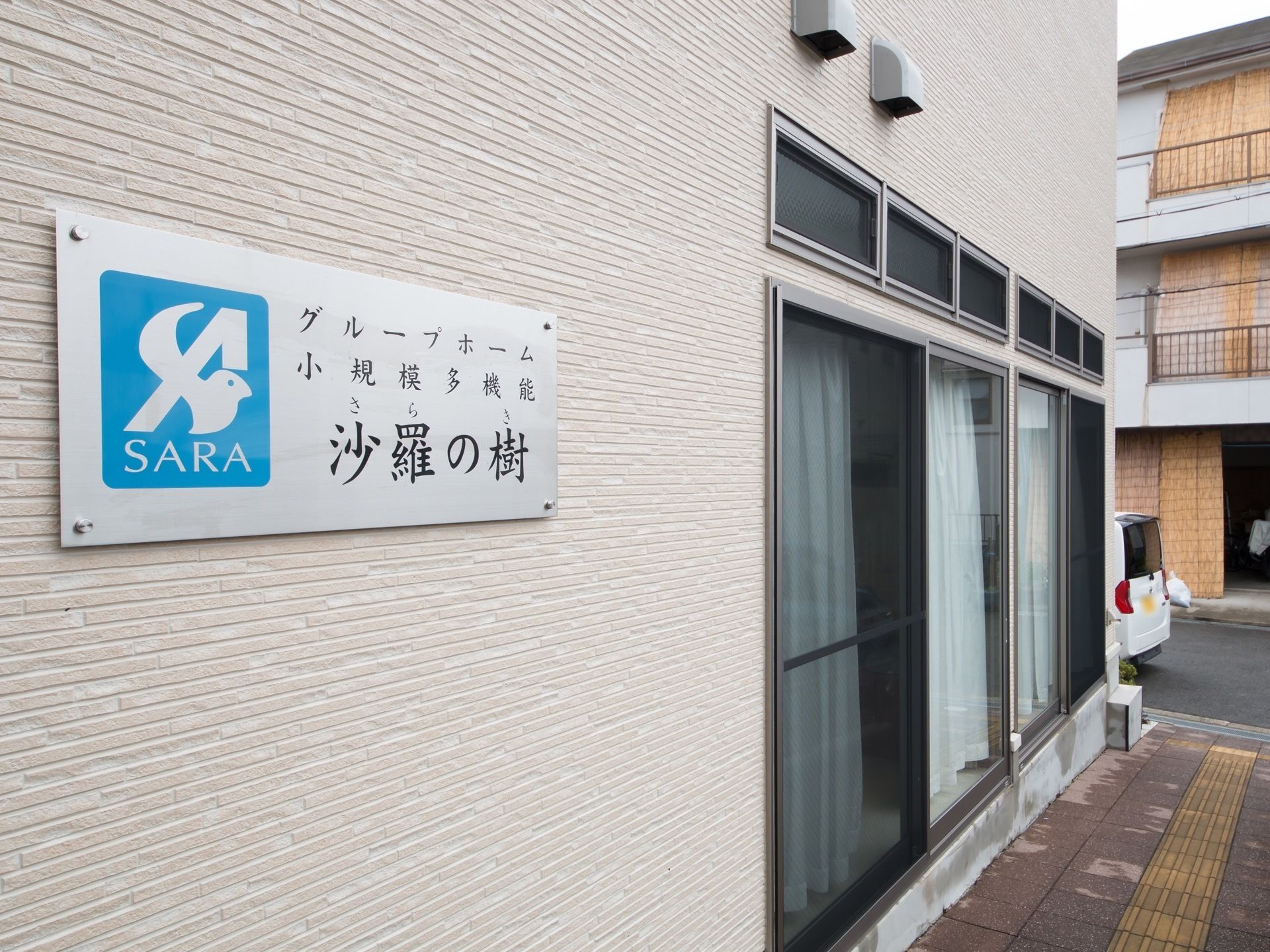 施設の白い外壁には施設名が記された看板が取りつけられており、大きな窓が設けられている。建物に沿って誘導ブロックが設置されている。