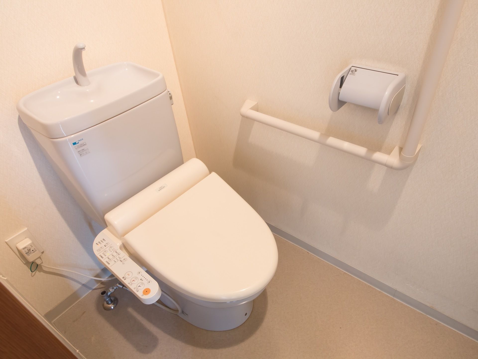 居室に備わった白ベースの清潔感のあるトイレである。立ち座りの際には、壁に設置されたＬ字型の手すりをつかむことができ安心である。