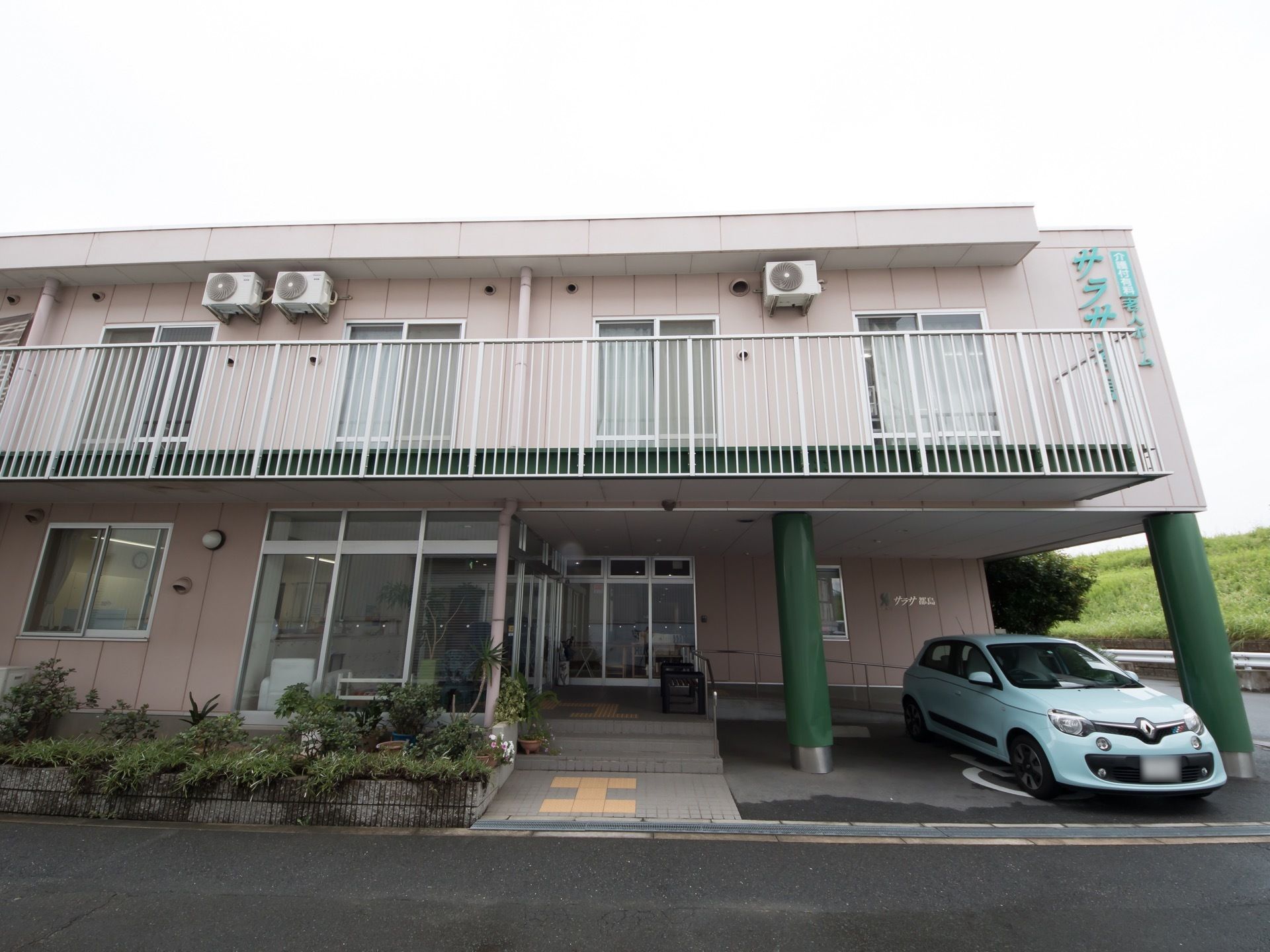 柔らかい配色の２階建て施設の外観である。２階の各居室には、見通しの良い柵のついたベランダが設置されている。