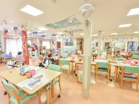 広々とした空間に、沢山のテーブルや椅子が配置されている。複数の入居者様が自由に過ごしている。柱には扇風機が取り付けられている。
