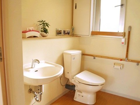 室内にはタンク付きのトイレと手洗い場があり、壁には長い手すりが取りつけられている。棚には、小さな植物が置かれている。