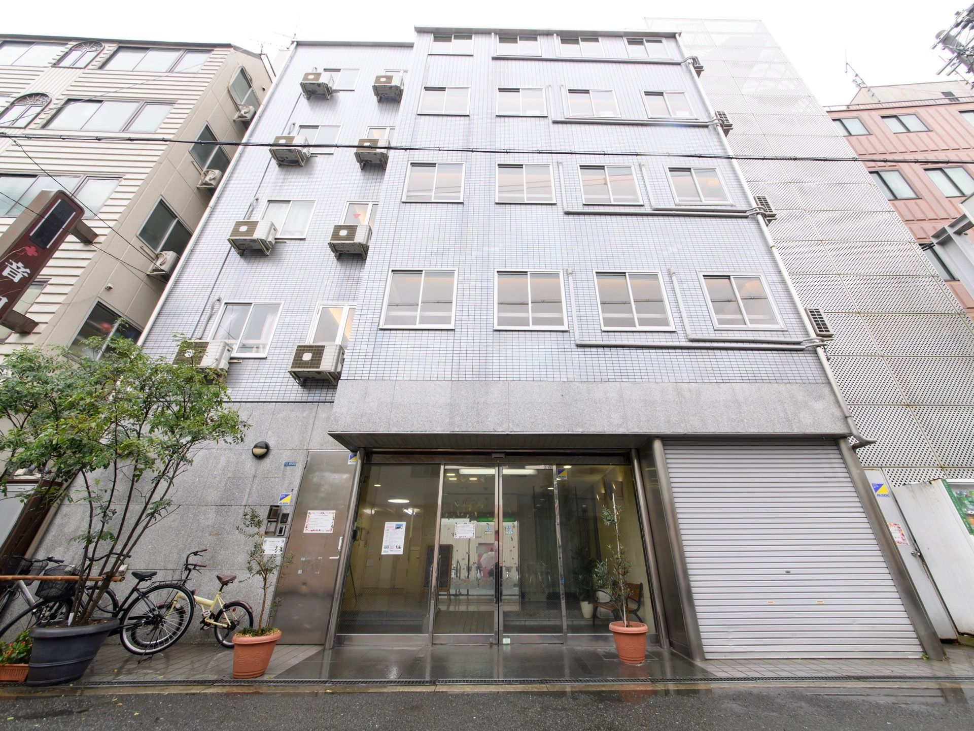 うすいグレーの壁で５階建てである。玄関ドアの左には駐輪スペースがあり、その前に大きな木が植えられている。
