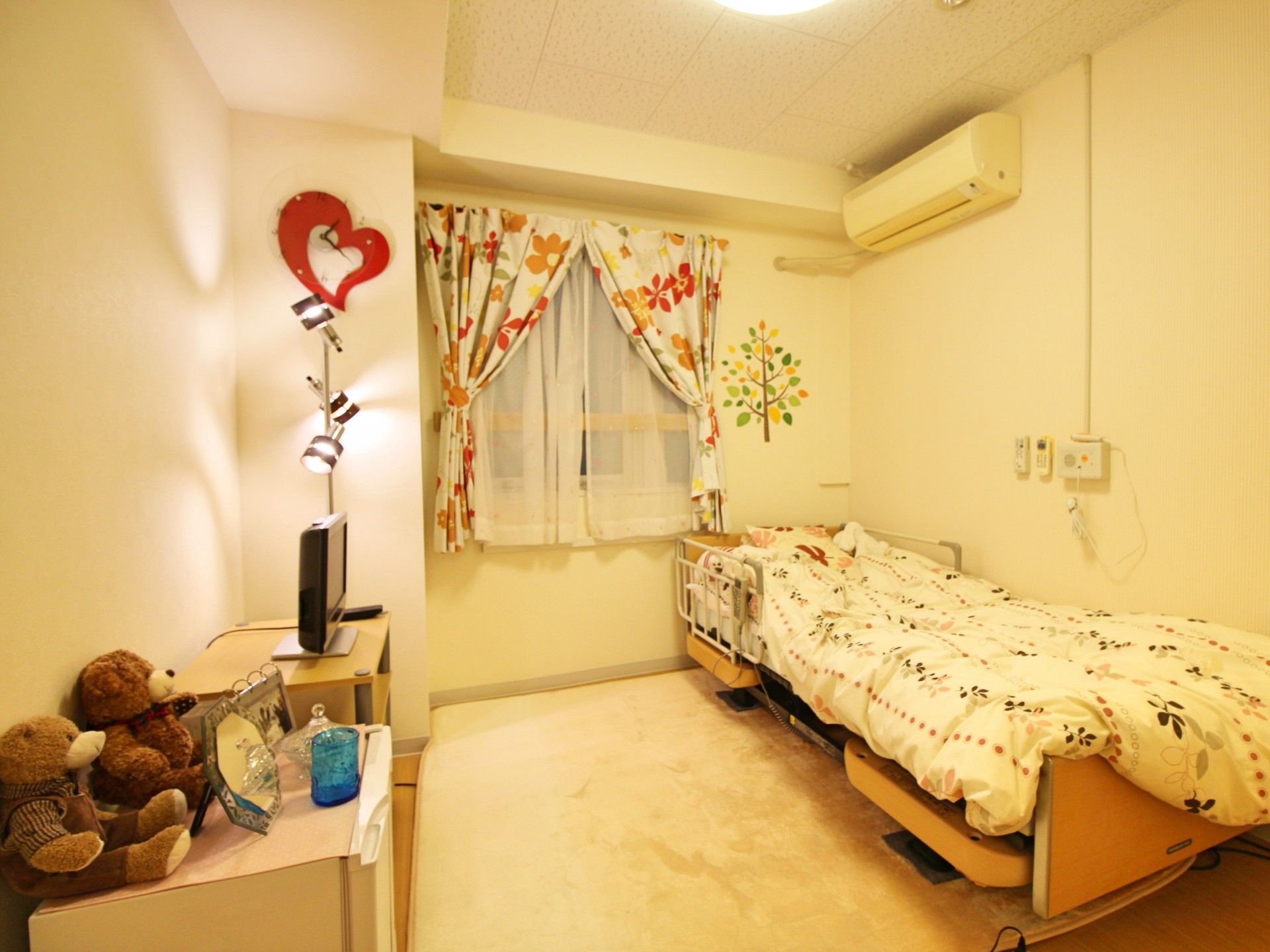 居室には一人で生活するのに十分な広さがあり、花柄のカーテンや布団、収納棚などがある。壁には、エアコンが設置されている。