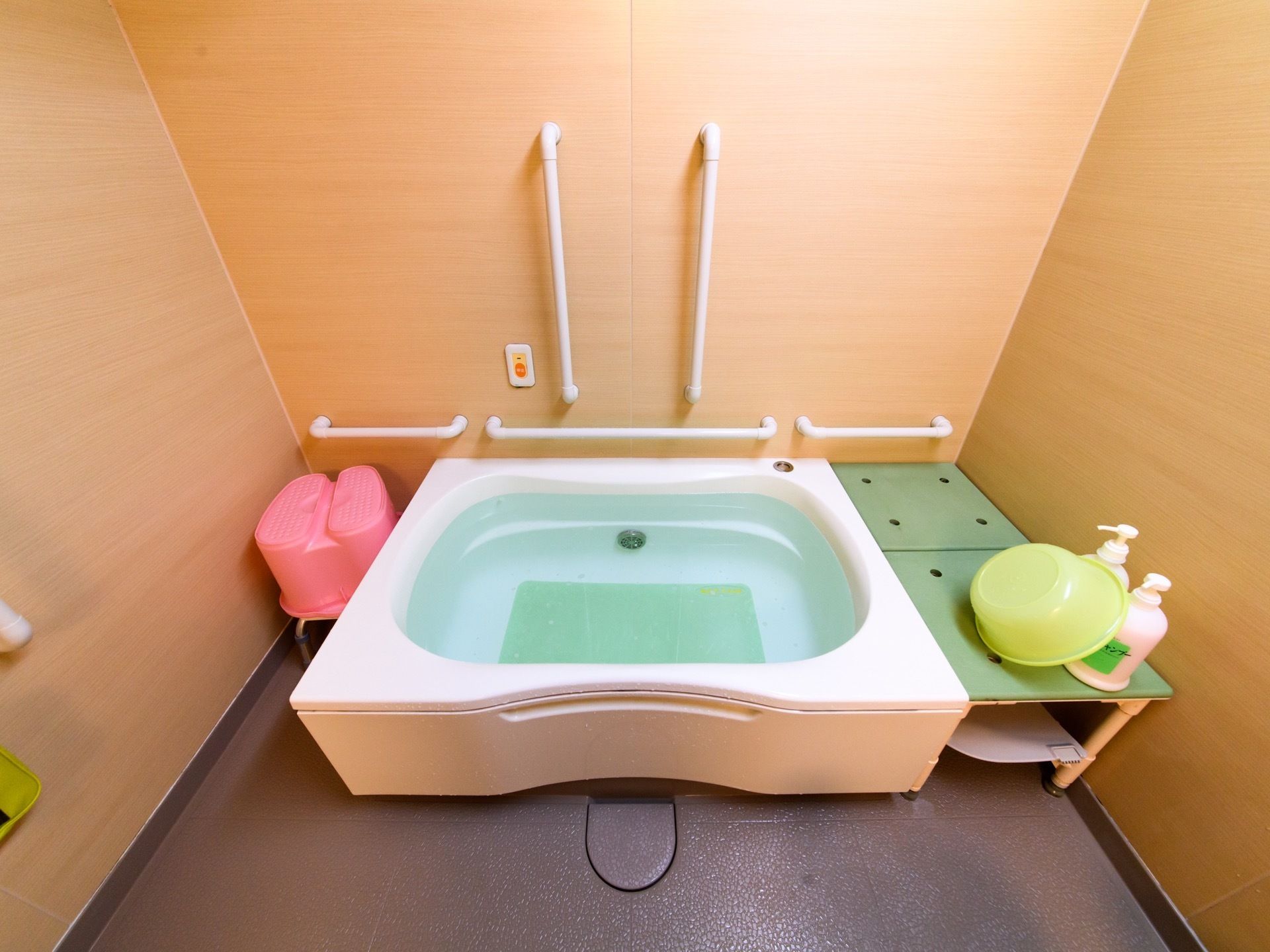 木目調の壁の明るい浴室である。壁の要所には手すりが設置されており、浴槽内には滑り込み防止用のマットが敷かれている。