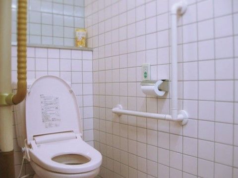 トイレはタイル張りになっている。Ｌ型手すりと呼び出しボダンが壁に設置されている。便器の後ろには、備品を置ける棚がある。