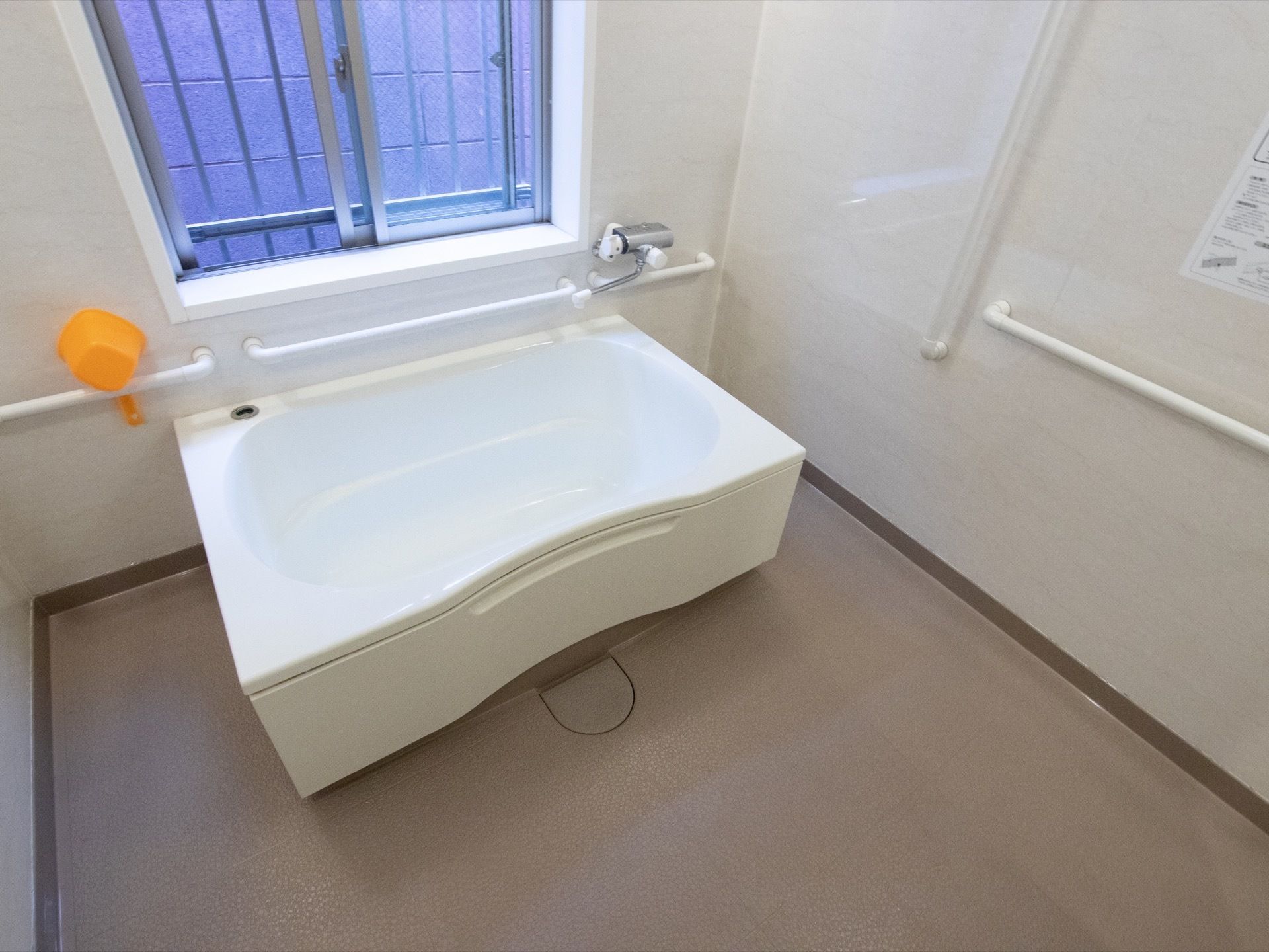 白色のシンプルなバスタブが置かれている。窓があるので換気が良く、壁には手すりが複数設置されている。手桶が用意されている。