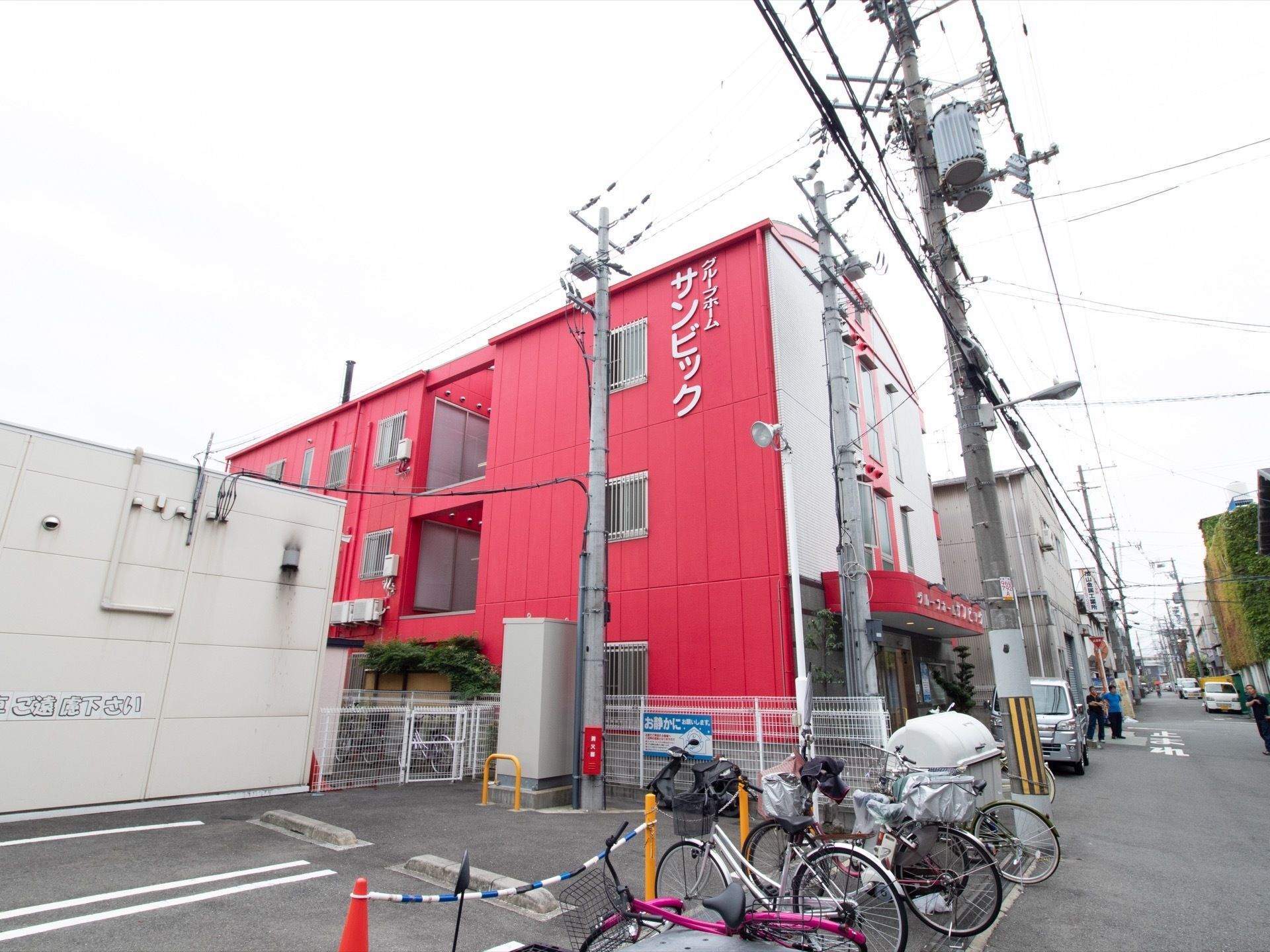 赤い外壁の３階建てのグループホームである。窓には白いフェンスが取り付けられており、玄関は屋根付きとなっている。