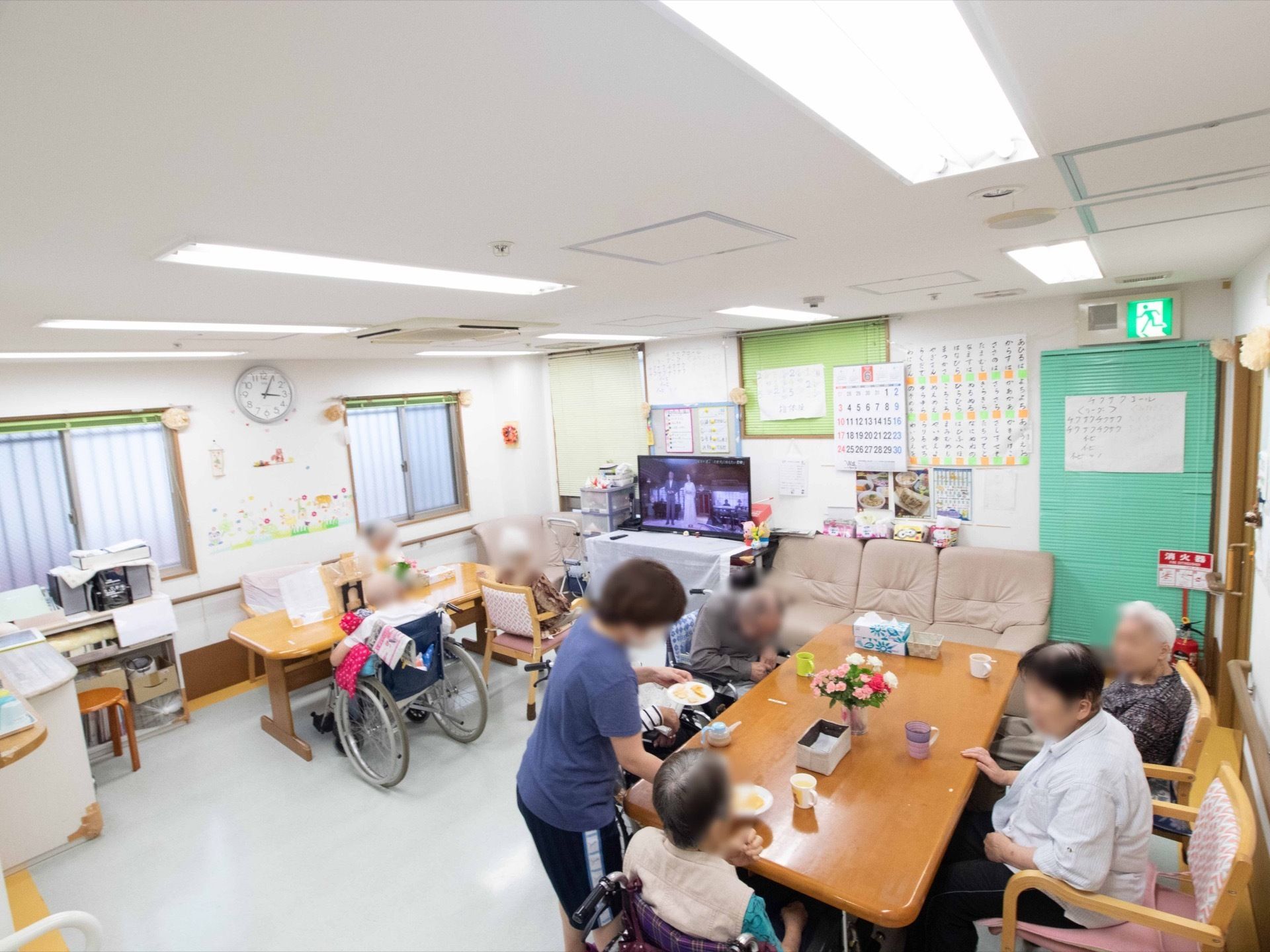 入居者様がテーブルを囲んでお茶を楽しんでいる。テレビの横にソファーが置いてあり、各窓にはブラインドが取り付けられている。