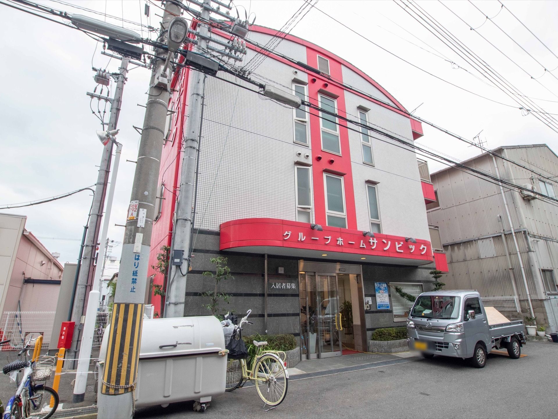 街中に建てられた施設で、グレーの外壁に赤色がアクセントとなっており、バリアフリーの玄関の上には施設名が大きく記載してある。