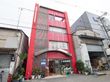 ４階建ての大きな赤い建物で、入口にはさまざまな種類の観葉植物が置かれている。窓には大きく入居者募集と書かれている。