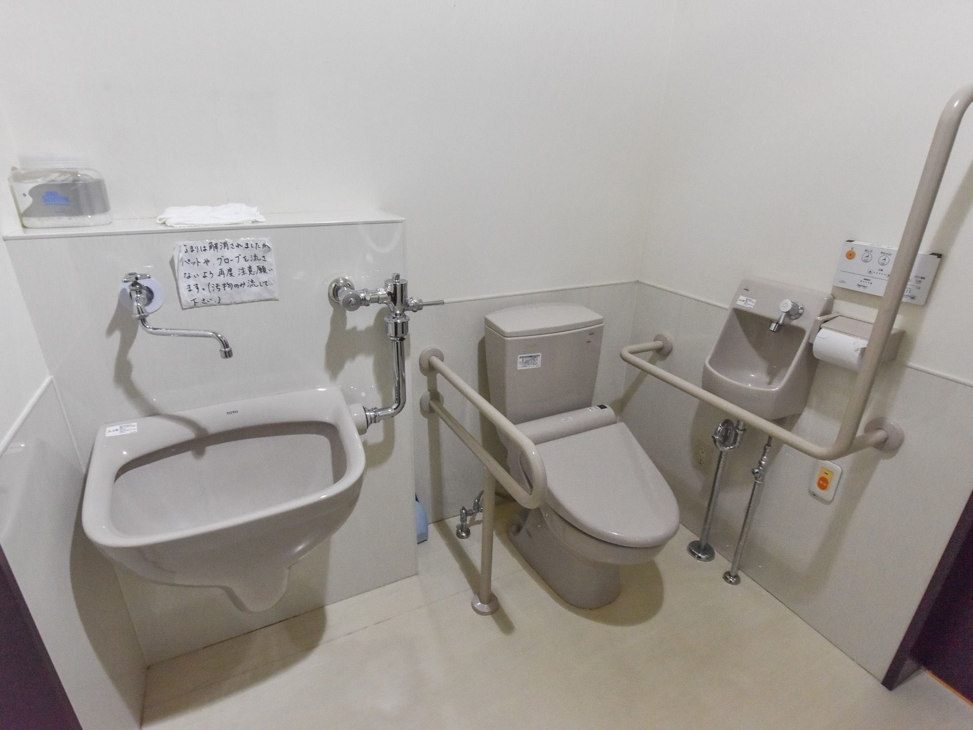 白とベージュの配色で清潔感のある空間である。便器の両脇には手すりがあり、洗面台も広い。壁には非常ボタンと温水洗浄便座のリモコンがある。