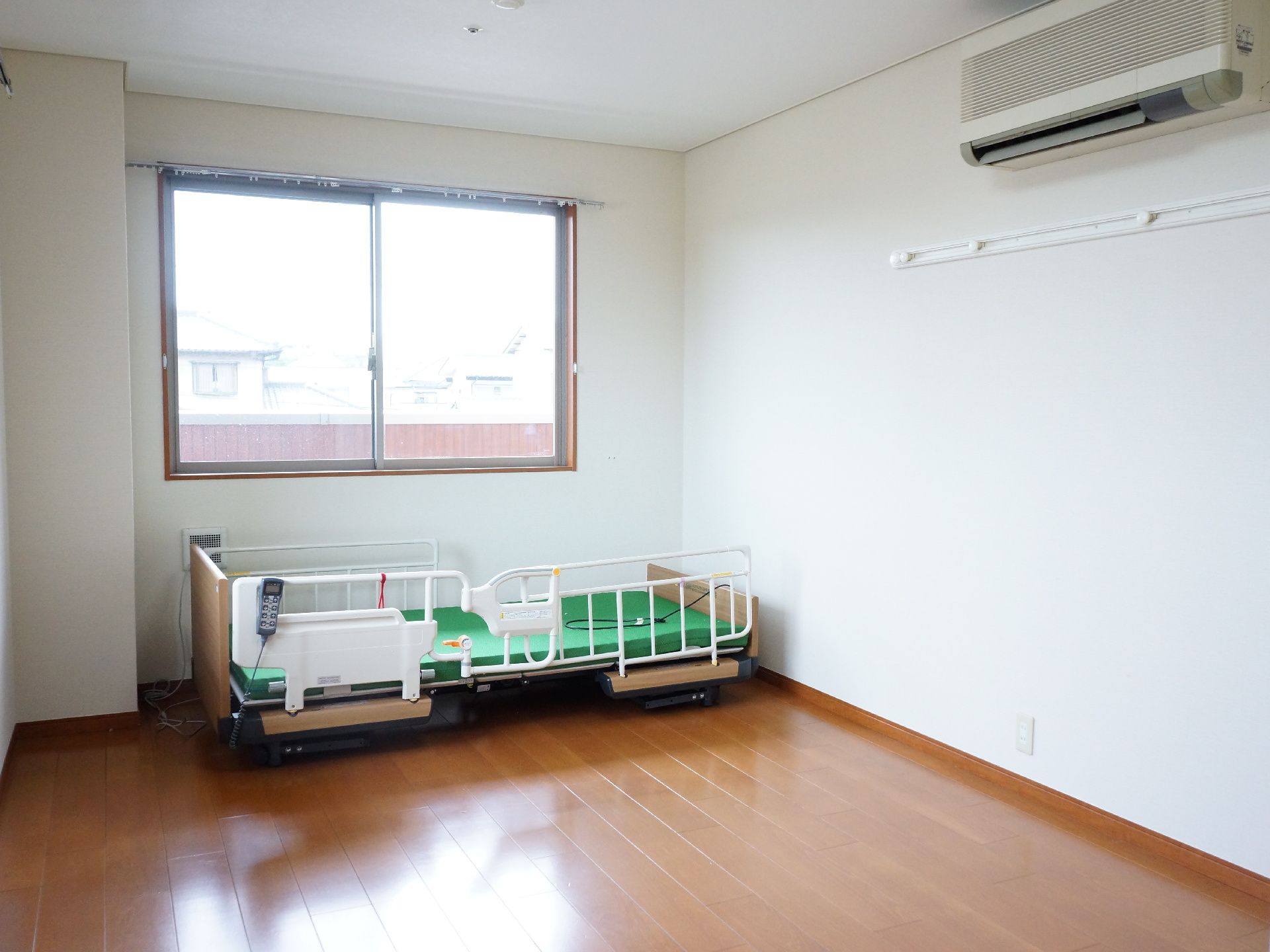 白色の天井と壁に茶色の床、正面と横側には窓がある。壁際には、介護用のベッドが置かれ、天井下にはエアコンが設置されている。