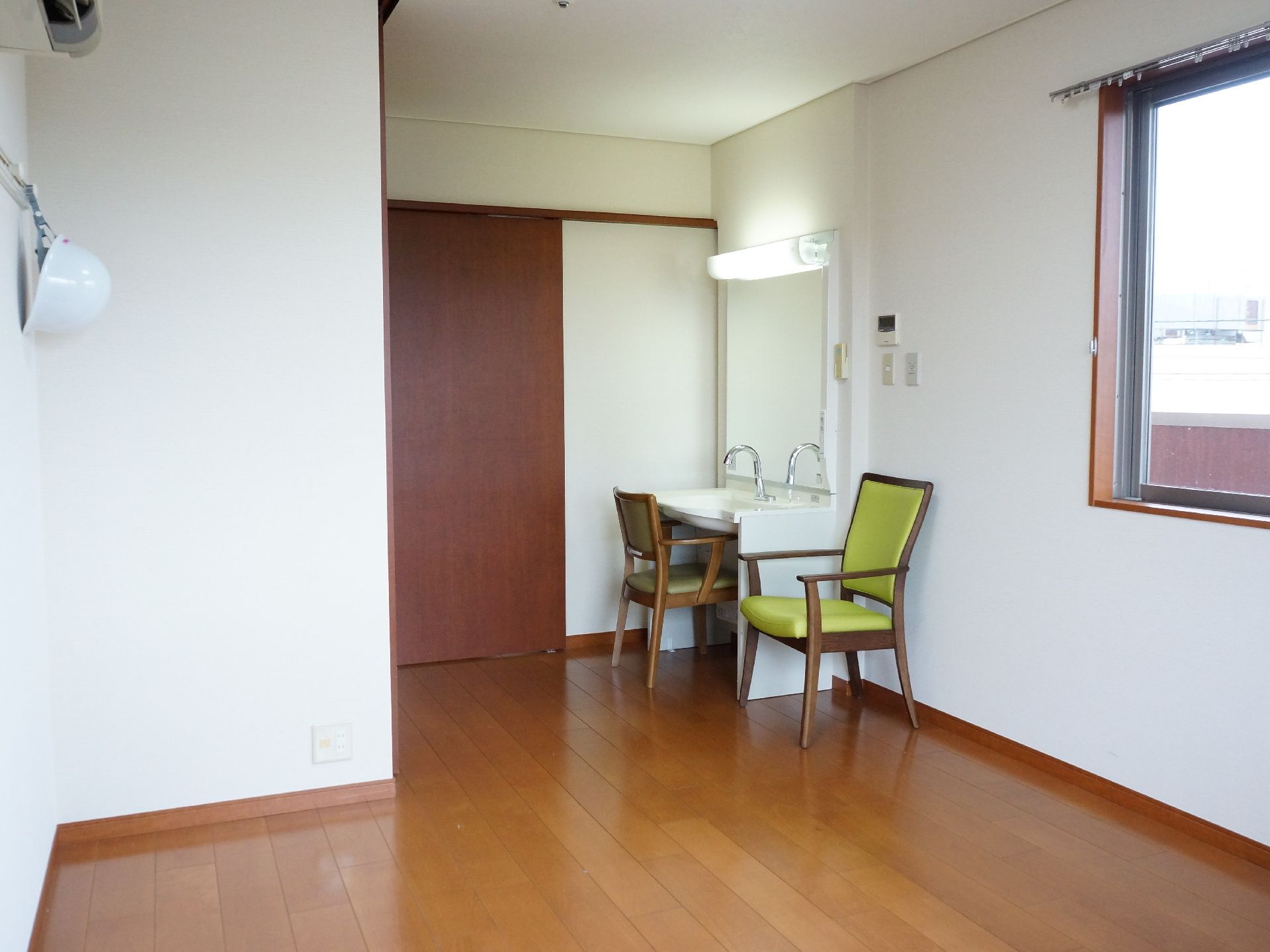 茶色の引き戸のドアから入って左側には、洗面台が設置してある。正面には鏡があり、その上には照明がある。そして、その前と横に椅子が置いてある。
