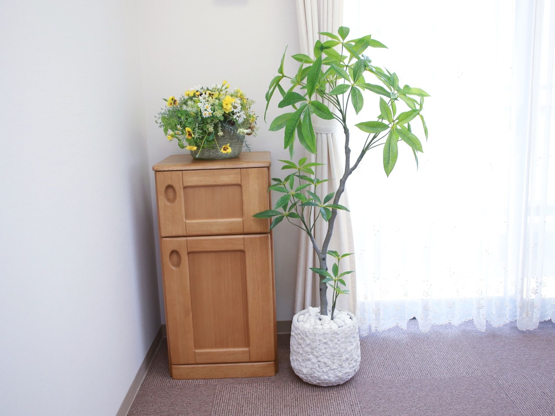 窓側の隅に、扉のついた木製家具が置かれており、その上に花が飾られている。隣に、観葉植物パキラの鉢植えが置かれている。