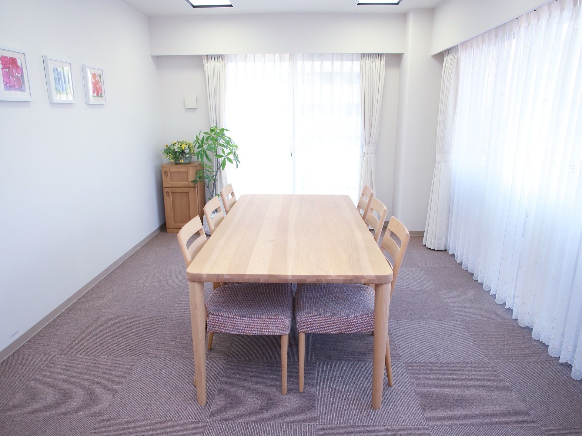 窓が多い明るい個室の中央に白木のテーブルと椅子６脚が置かれている。壁に絵画３点が並べて飾られており、部屋の隅に観葉植物がある。