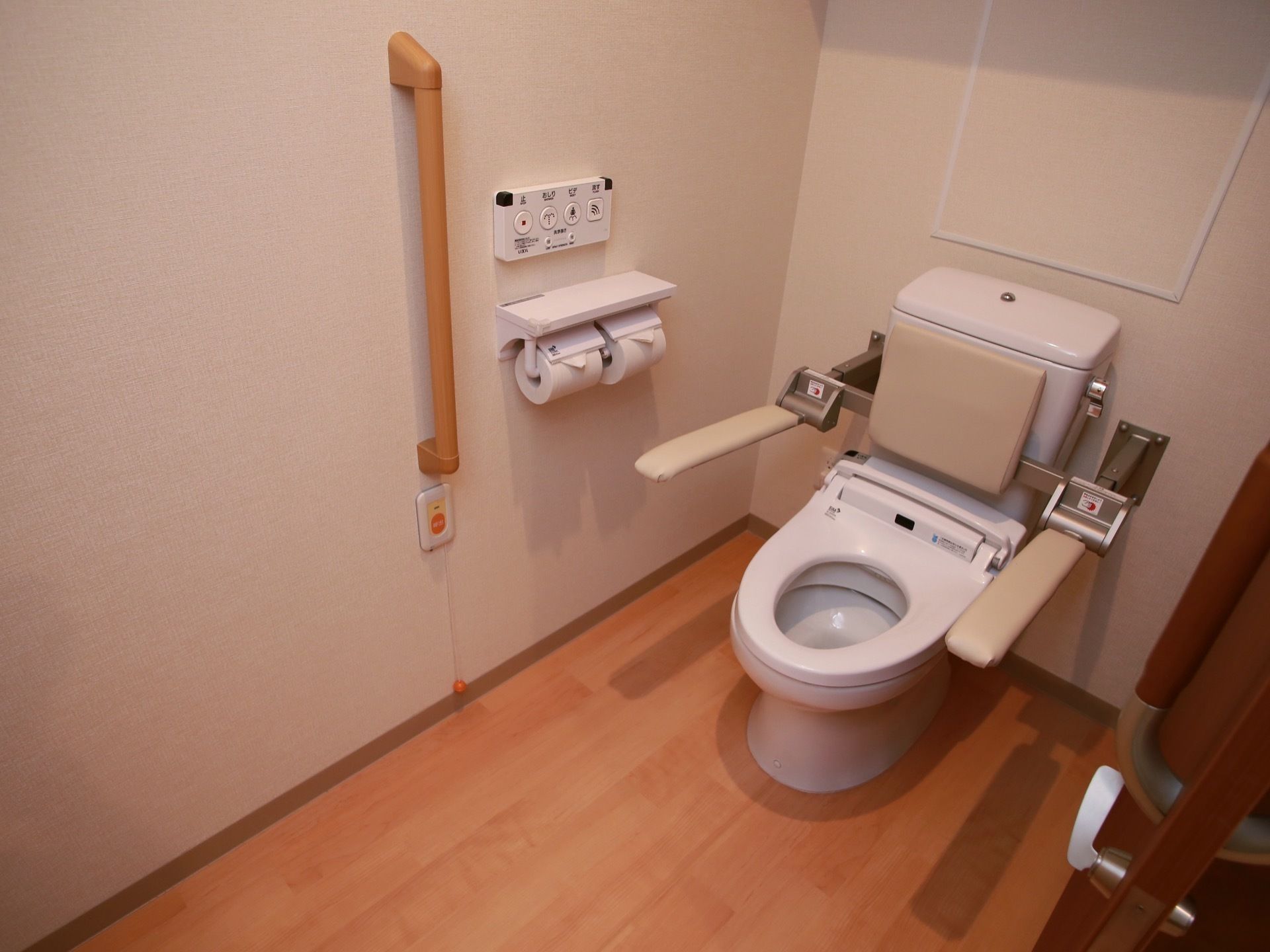 蓋のない便器に、背もたれとアームレストが取りつけられている。手すりの下にナースコールが設置されている。