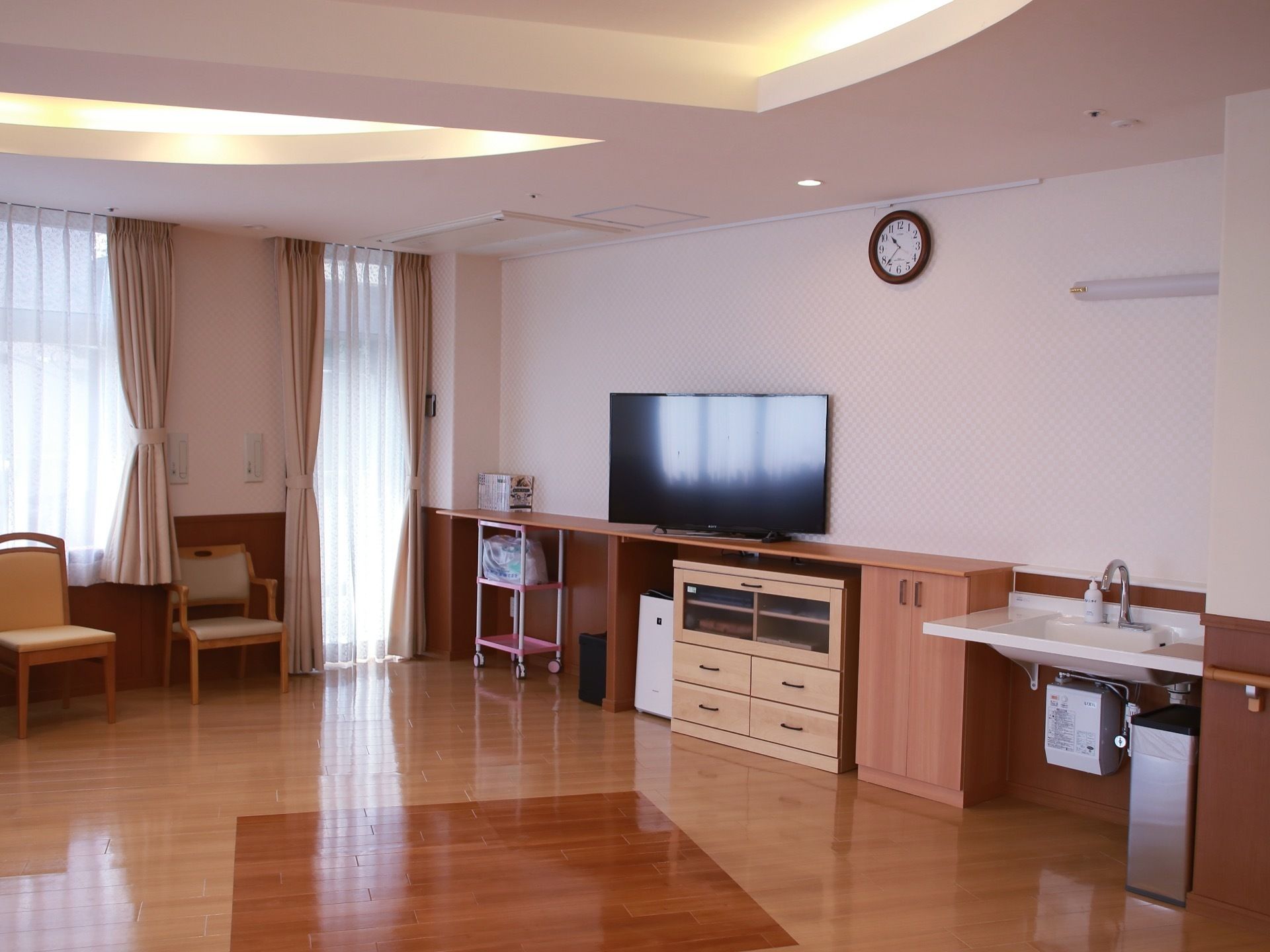 壁際の棚の上に大型液晶テレビが設置されている。幅の広い洗面台や、クッションが施された椅子などが見える。
